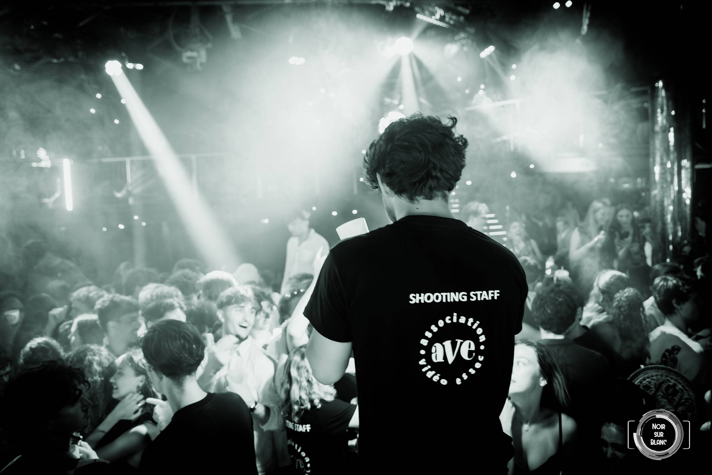 Photo en noir et blanc : une personne en tee-shirt marqué Shooting Staff, de dos, fait face à la foule éclairée par les projecteurs.
