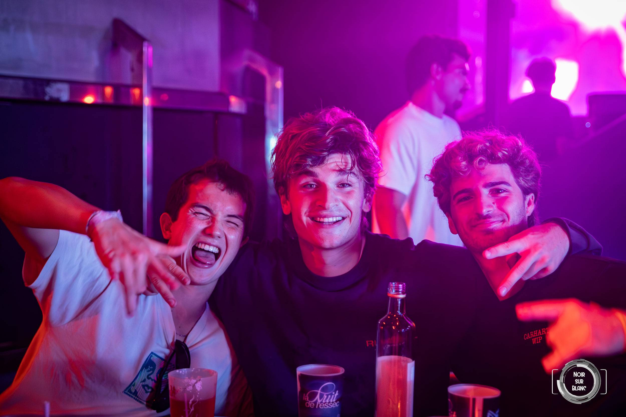 Trois jeunes hommes se tiennent par les épaules en riant, sous une lumière magenta, une bouteille et des gobelets posés devant eux.