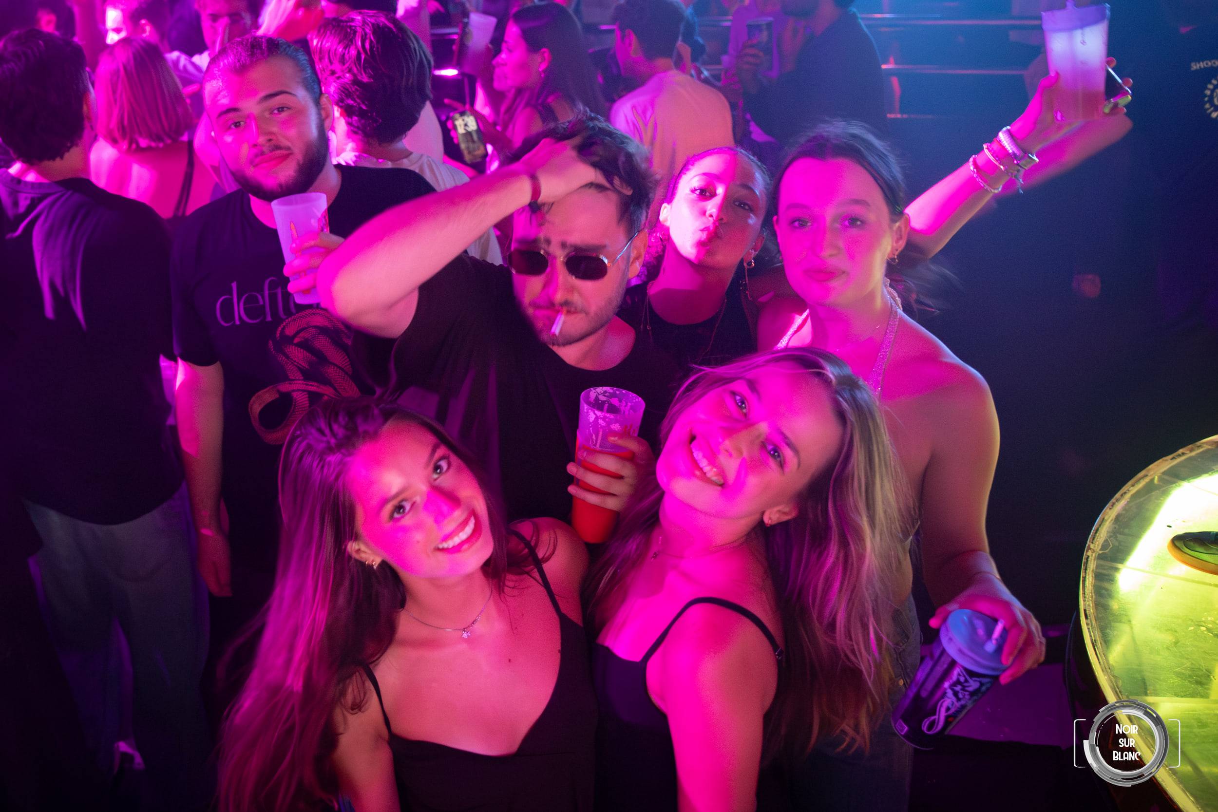 Un groupe pose serré sous une lumière rose, l'un d'eux brandissant une bouteille lumineuse au-dessus des têtes.
