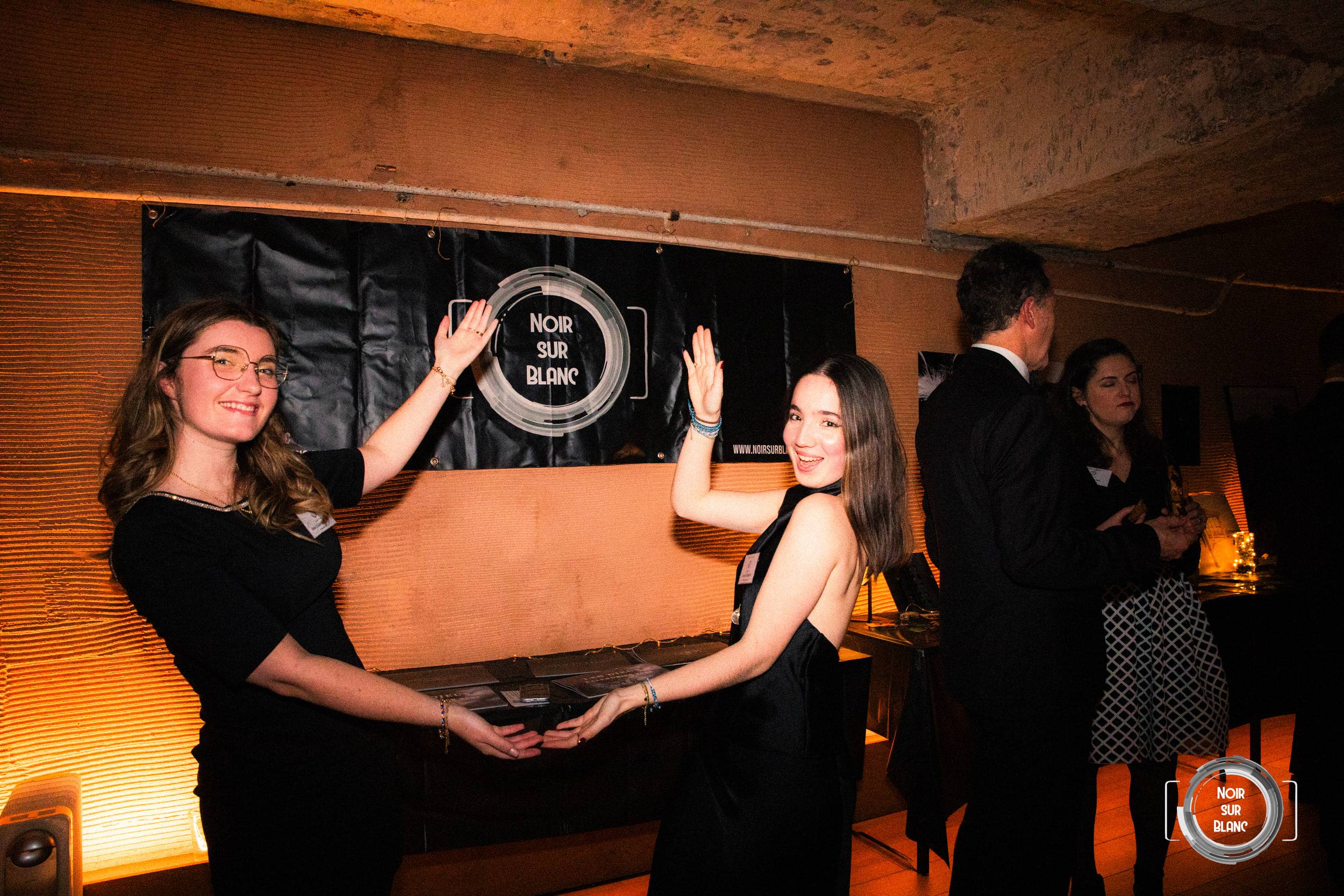Deux invitées en robe noire présentent un plateau à l'objectif sous une banderole noire au logo de Noir sur Blanc.
