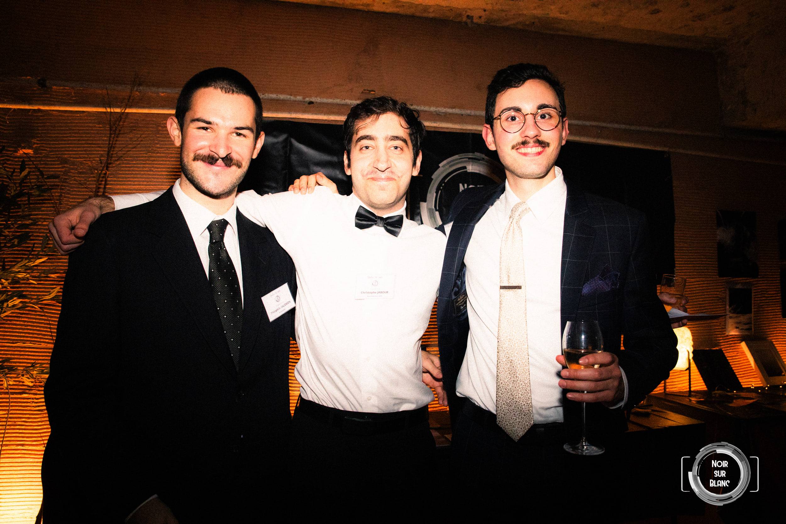 Trois invités en smoking, bras sur les épaules, posent verre à la main ; celui du milieu est en chemise blanche et nœud papillon noir.