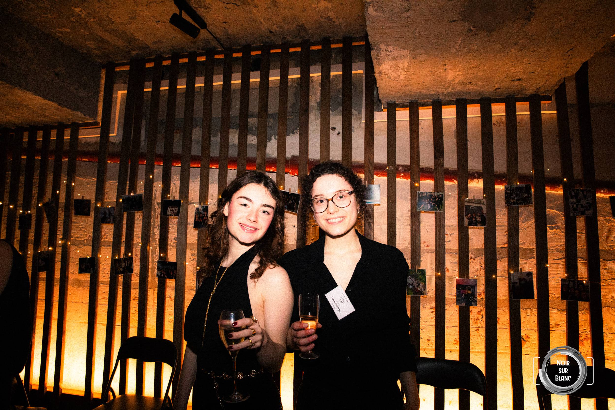 Deux invitées en noir, verre à la main, posent devant une claustra de bois éclairée par en dessous.