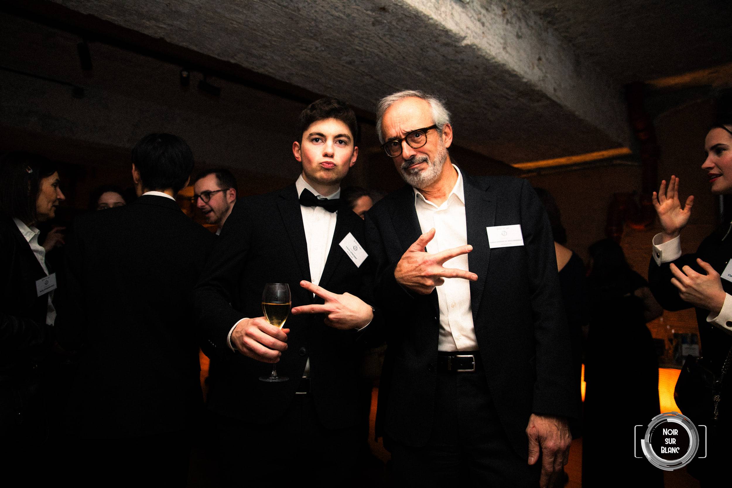 Un étudiant en smoking et un invité plus âgé en chemise blanche se saluent, verre à la main, au milieu de la soirée.