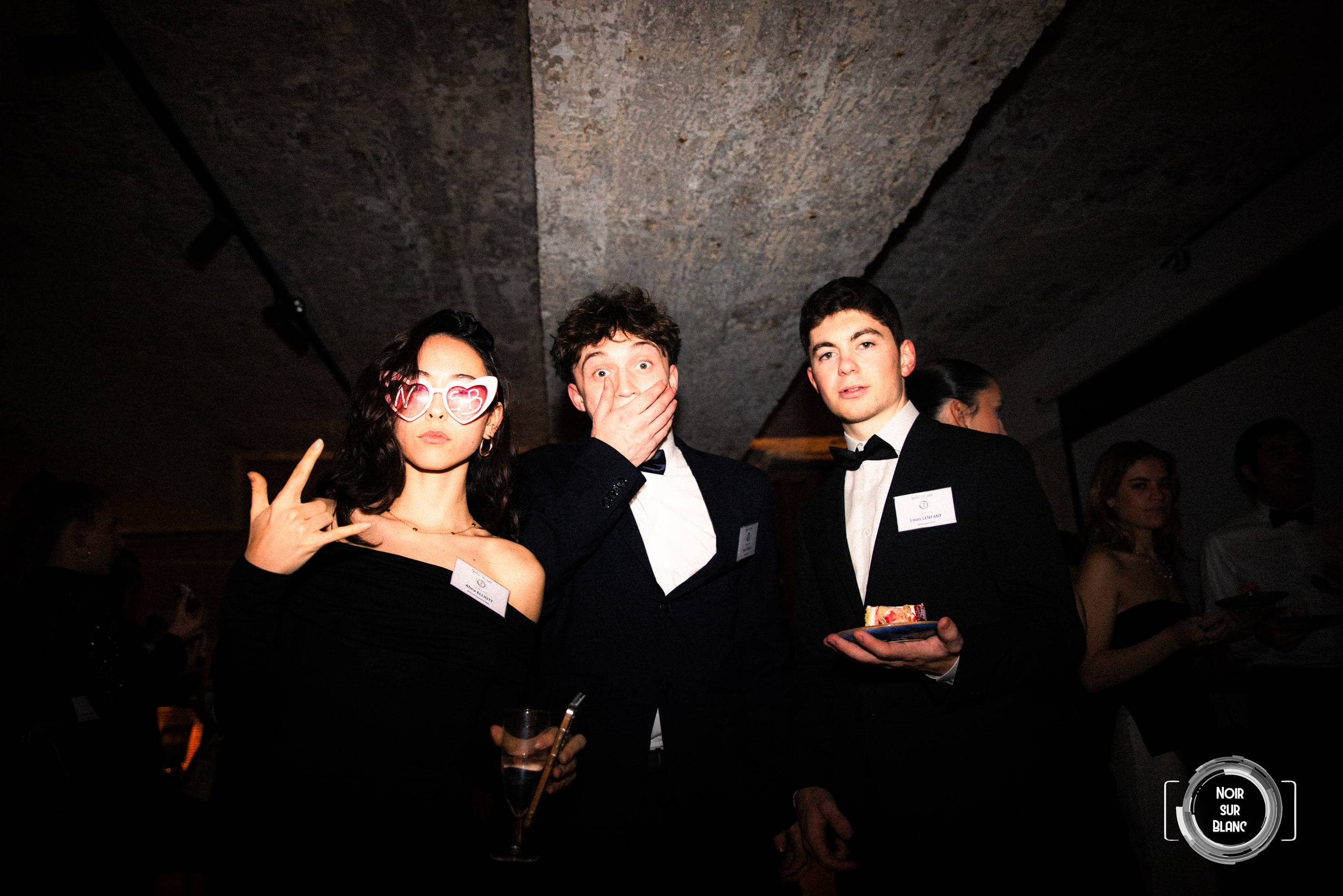 Trois invités en tenue de soirée face à l'objectif : une jeune femme porte des lunettes en forme de cœur marquées NSB, son voisin plaque une main sur sa bouche, le troisième tient une assiette de gâteau.