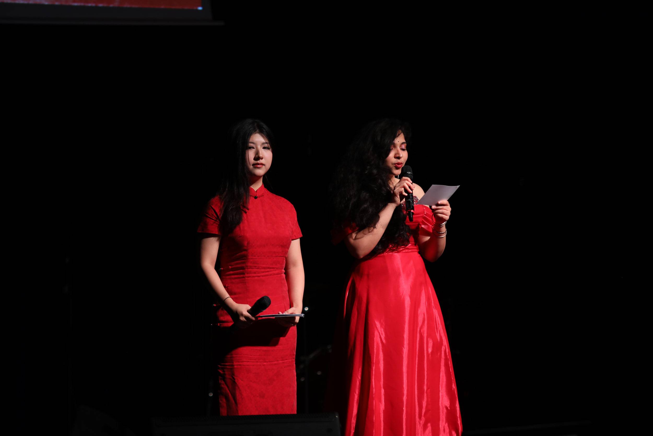 Deux présentatrices en robe rouge, l'une en qipao, se partagent le micro sur scène.