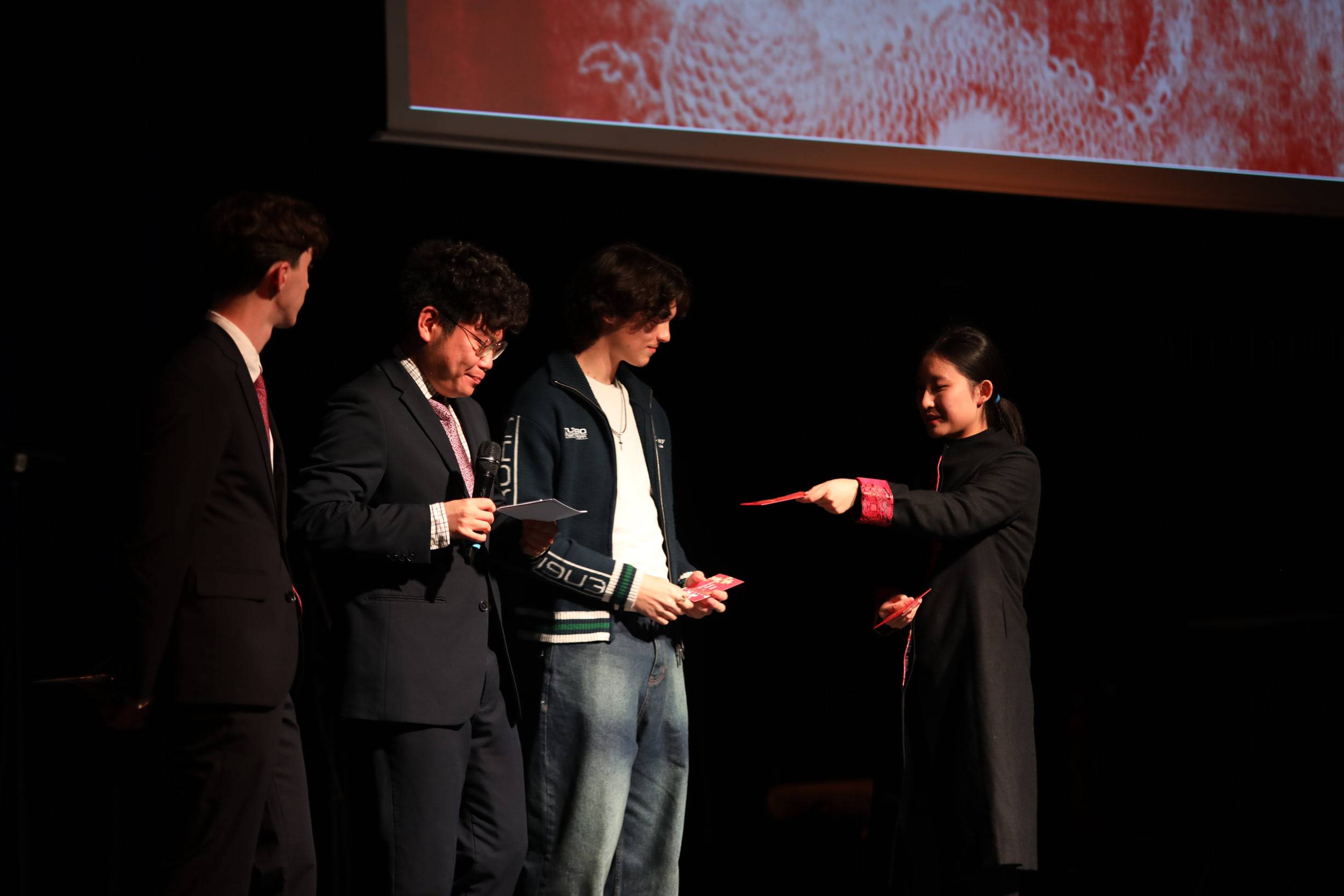 Sur scène, une organisatrice remet une enveloppe rouge à un étudiant sous le regard des présentateurs.