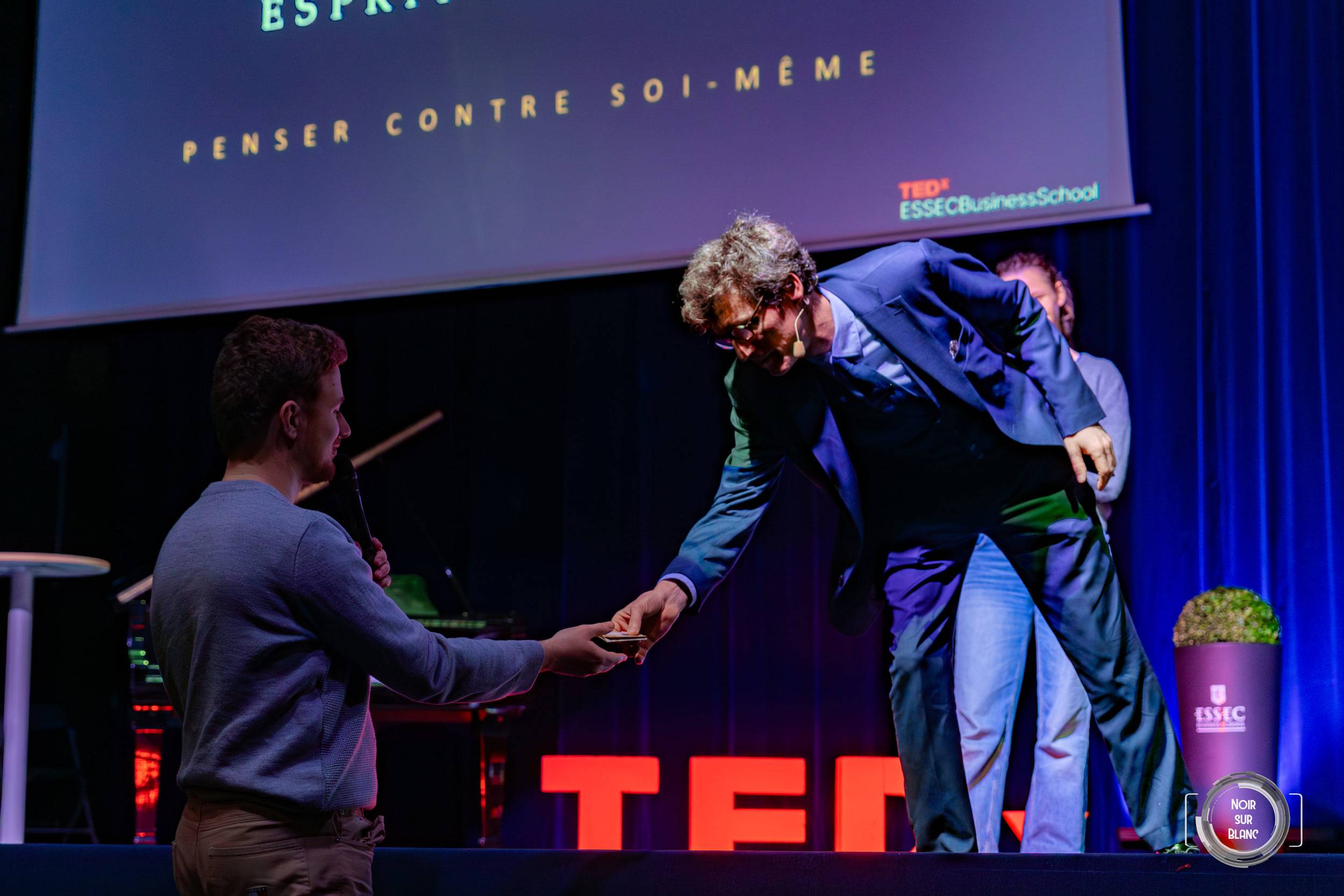 Deux hommes se serrent la main au bord de la scène, devant les lettres rouges du logo TEDx.
