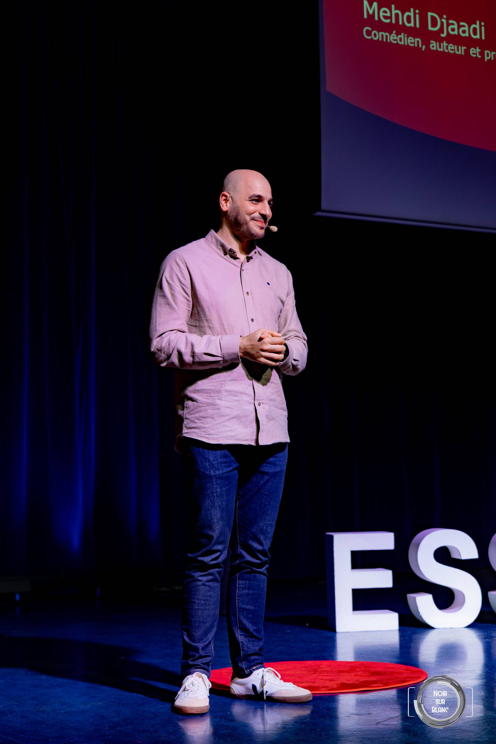 Un intervenant en chemise claire parle debout sur le tapis rond rouge d'une scène TEDx, les lettres ESSEC éclairées derrière lui.