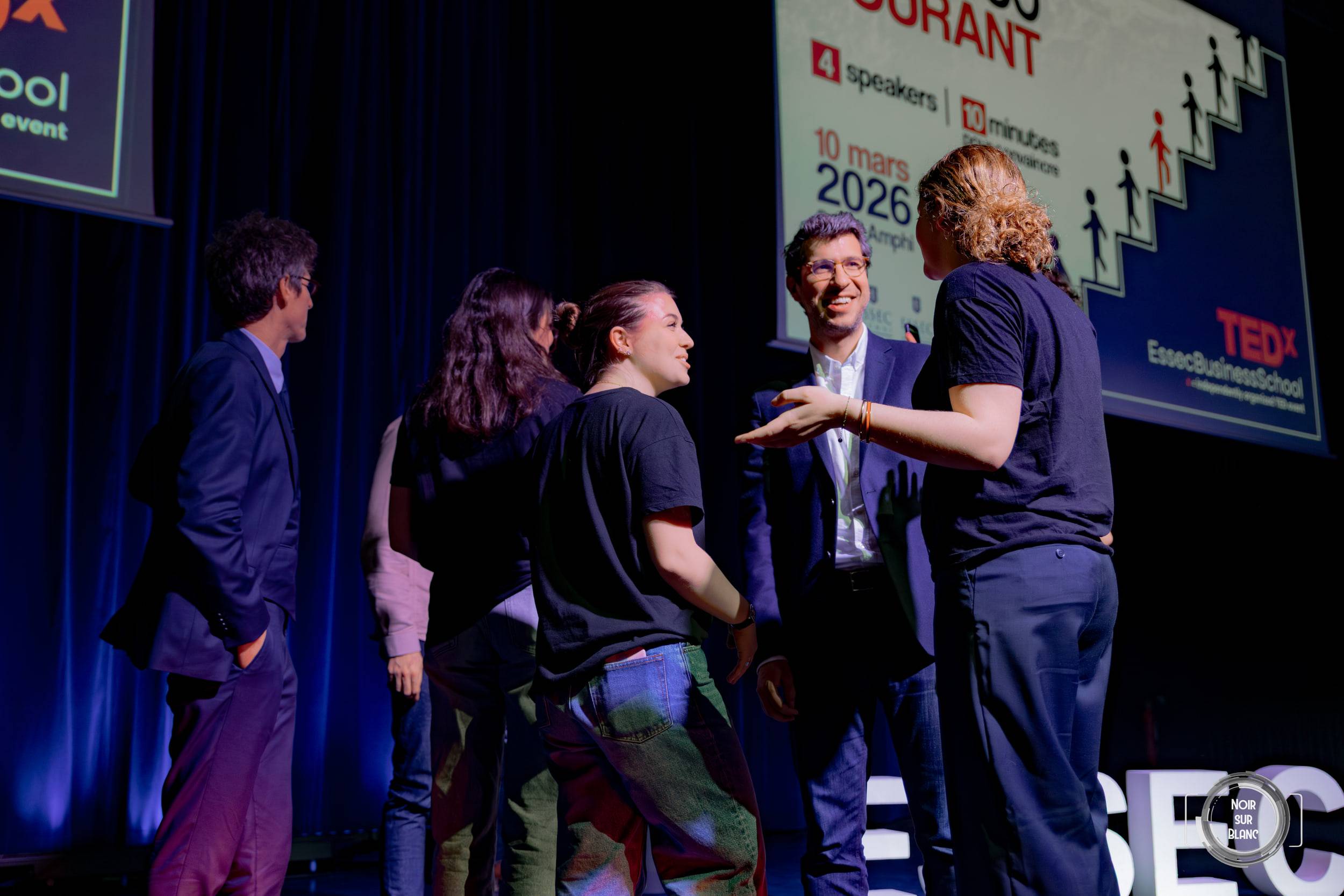 L'équipe organisatrice, en t-shirt noir, applaudit sur scène devant l'affiche de l'événement.