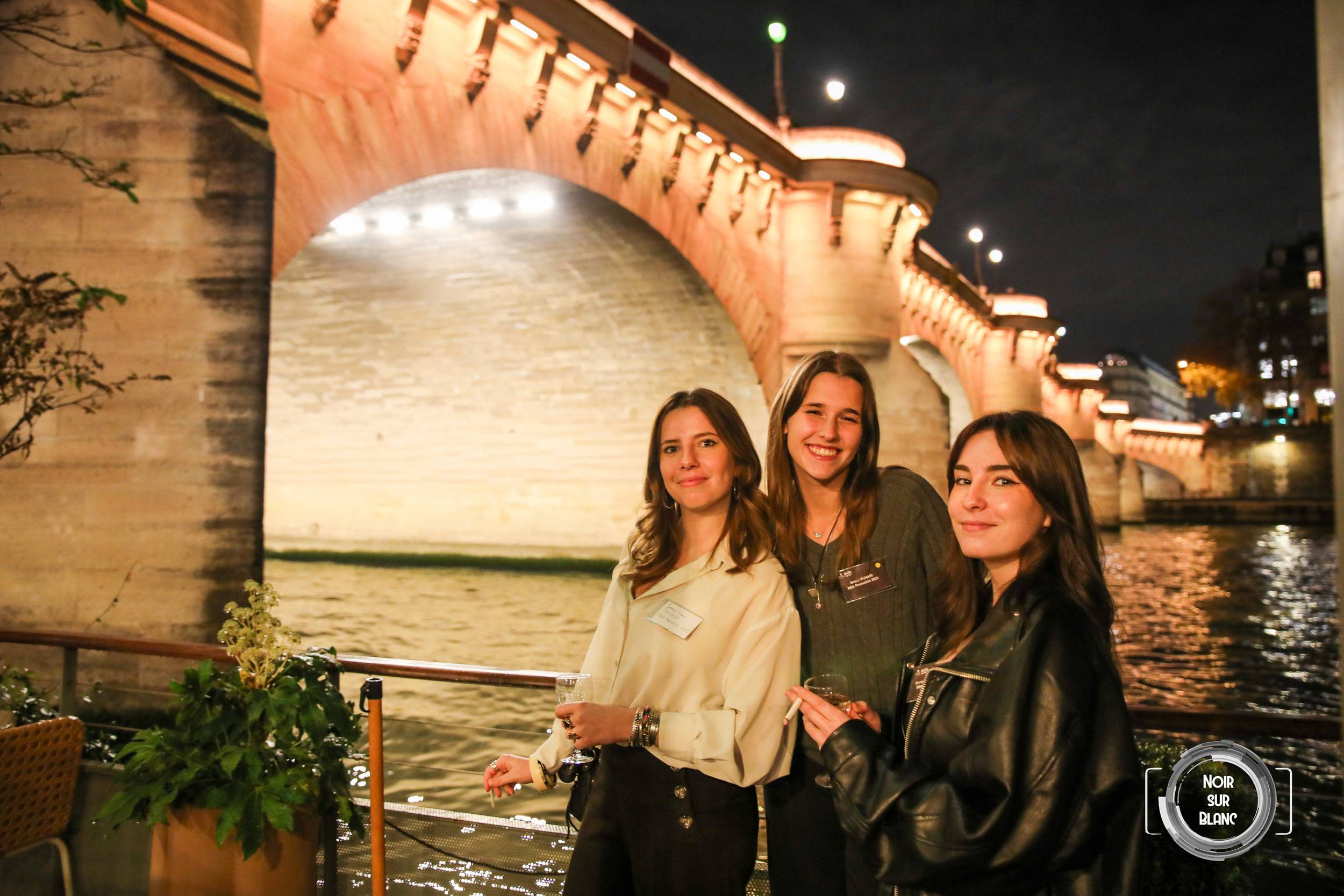 Trois invitées posent sur le pont d'une péniche, les arches éclairées d'un pont de Paris derrière elles.