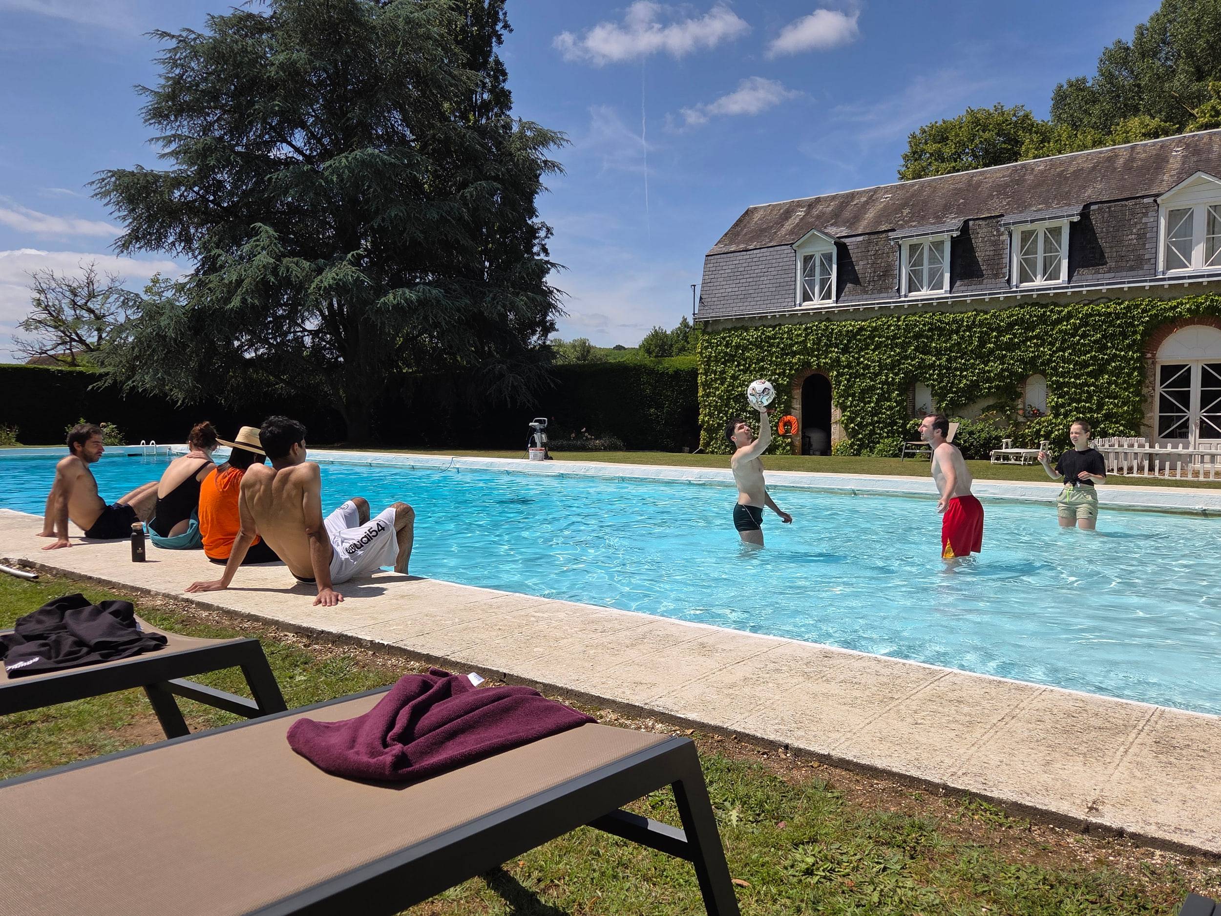 Vue large de la piscine devant une longère couverte de lierre : plusieurs personnes dans l'eau, d'autres sur les transats.