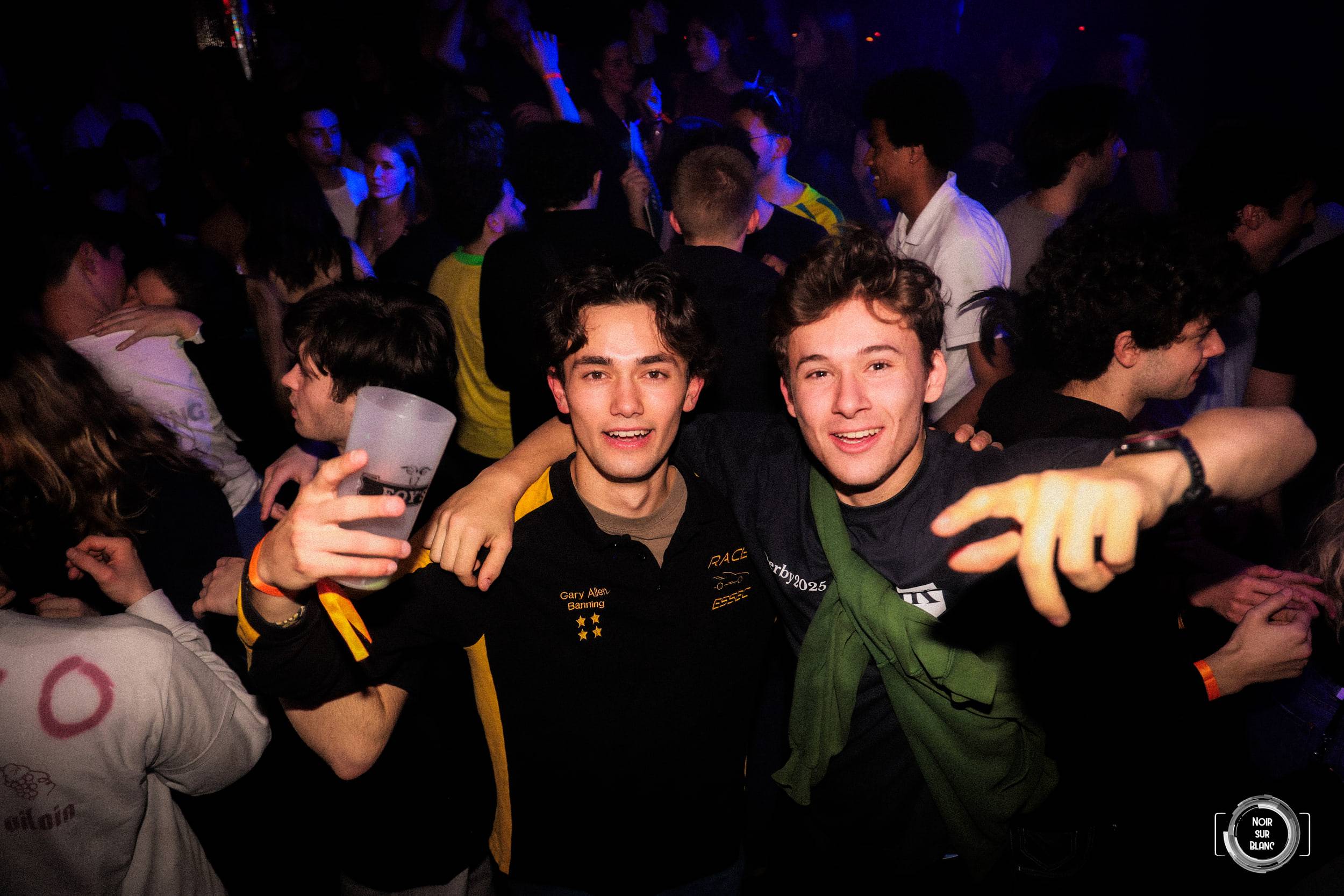 Deux étudiants tendent le bras vers l'objectif, verre en main, dans les lumières bleues de la salle.