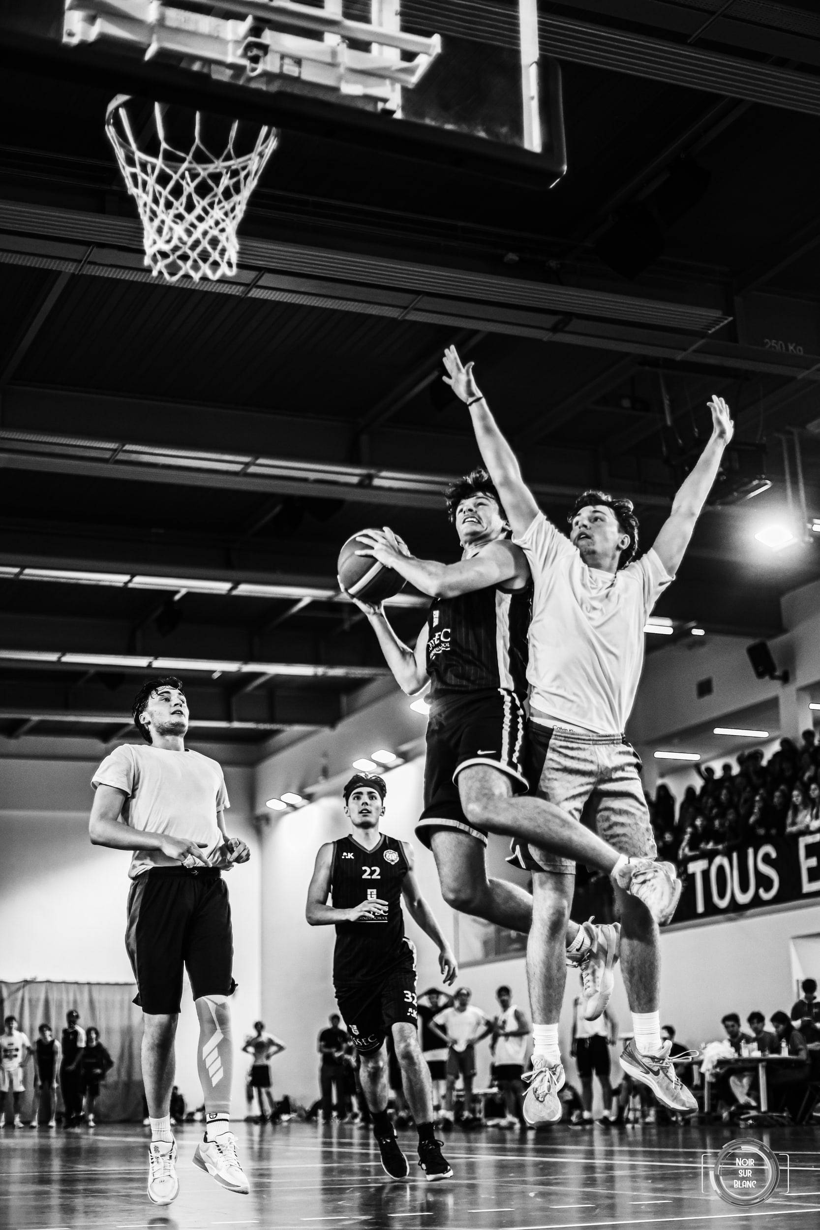 En noir et blanc, sous le panier vu en contre-plongée, deux joueurs s'élèvent au même instant pour disputer le ballon devant des tribunes pleines.
