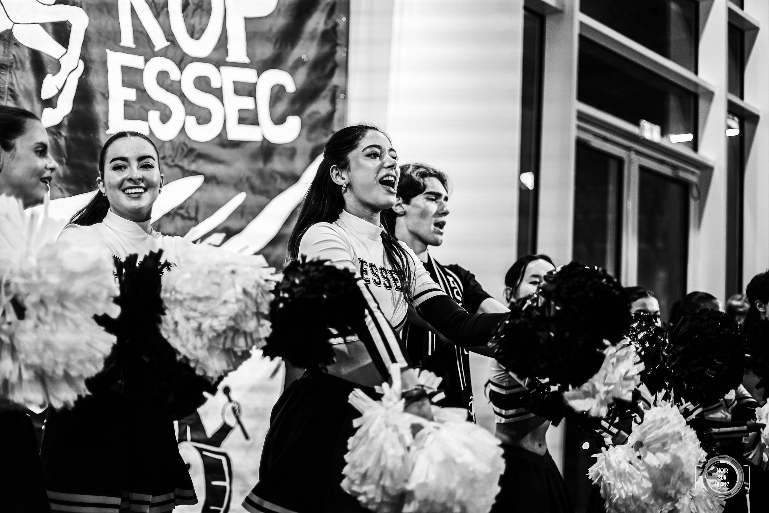 En noir et blanc, des pom-pom girls agitent leurs pompons devant une banderole marquée « KOP ESSEC », la bouche ouverte sur un chant.