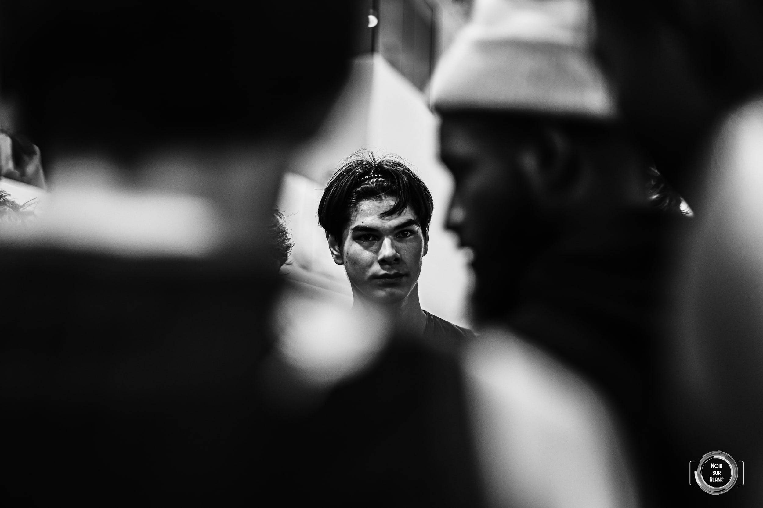 En noir et blanc, le visage d'un boxeur fixe l'objectif, encadré par deux silhouettes floues au premier plan.