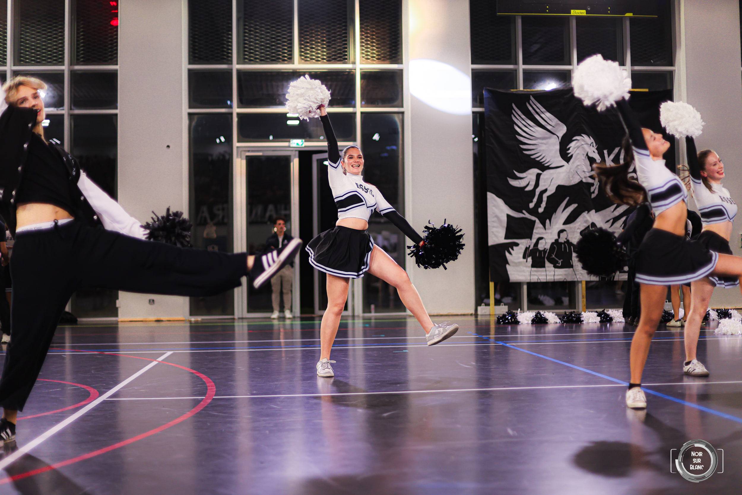 Au milieu du gymnase, une pom-pom girl est saisie en plein saut, jambes écartées et pompons tendus, devant le drapeau au pégase de l'école.