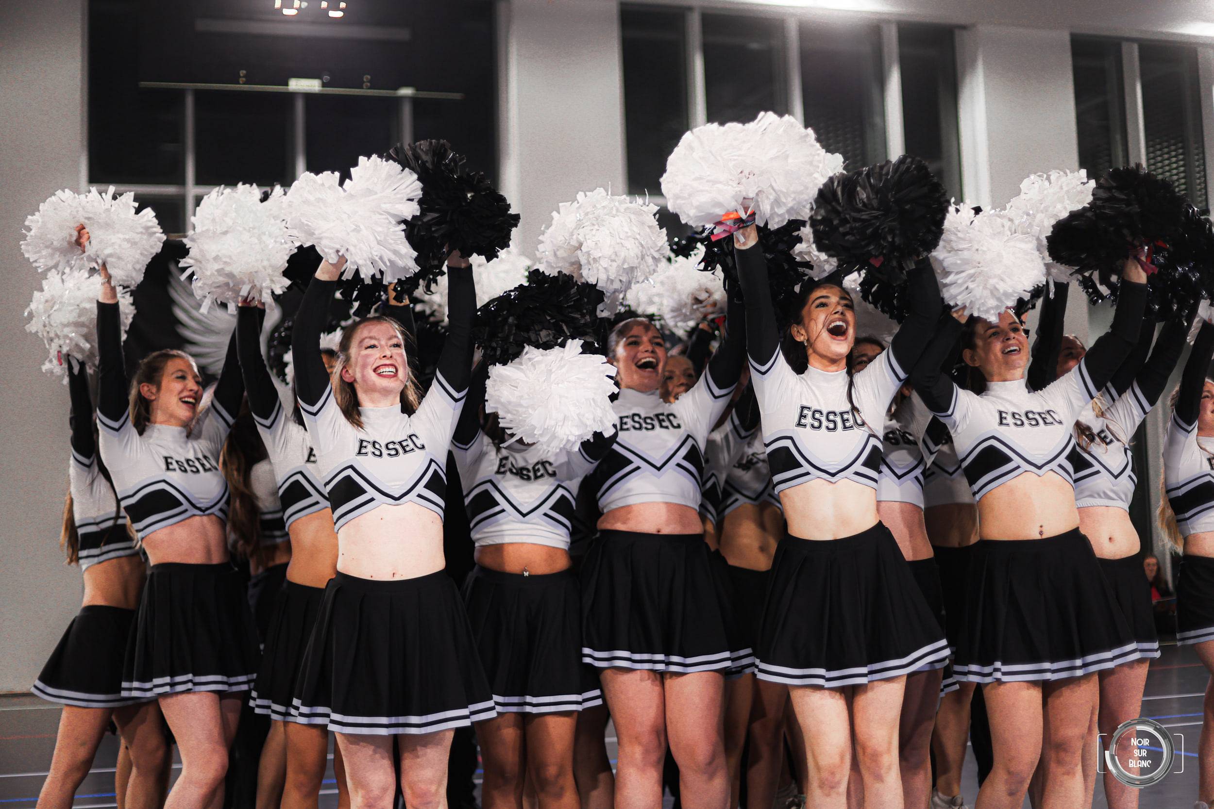 Une dizaine de pom-pom girls en tenue ESSEC noir et blanc lèvent leurs pompons au-dessus de leur tête, alignées face à la salle.