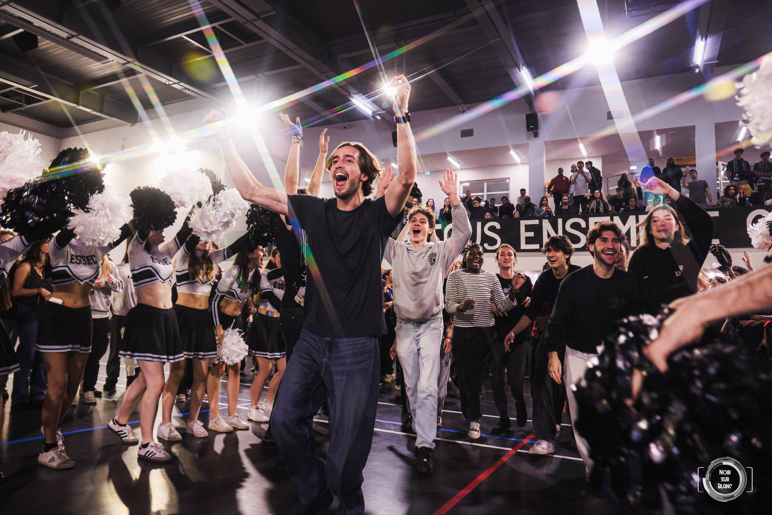 Bras levés au milieu du gymnase, un étudiant lance un chant devant les pom-pom girls et la tribune, dans les faisceaux des projecteurs.