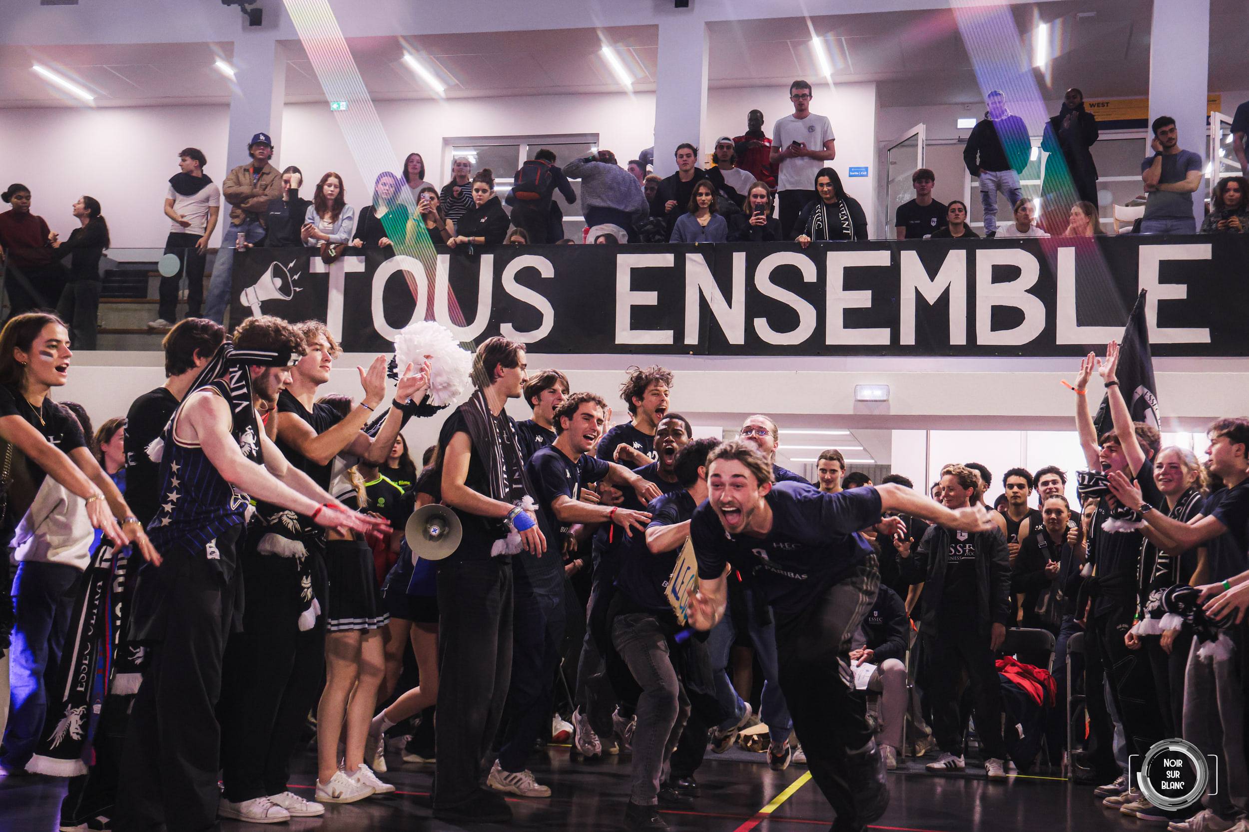 Sous une banderole noire marquée « TOUS ENSEMBLE », les supporters sautent et agitent des pompons dans le gymnase.