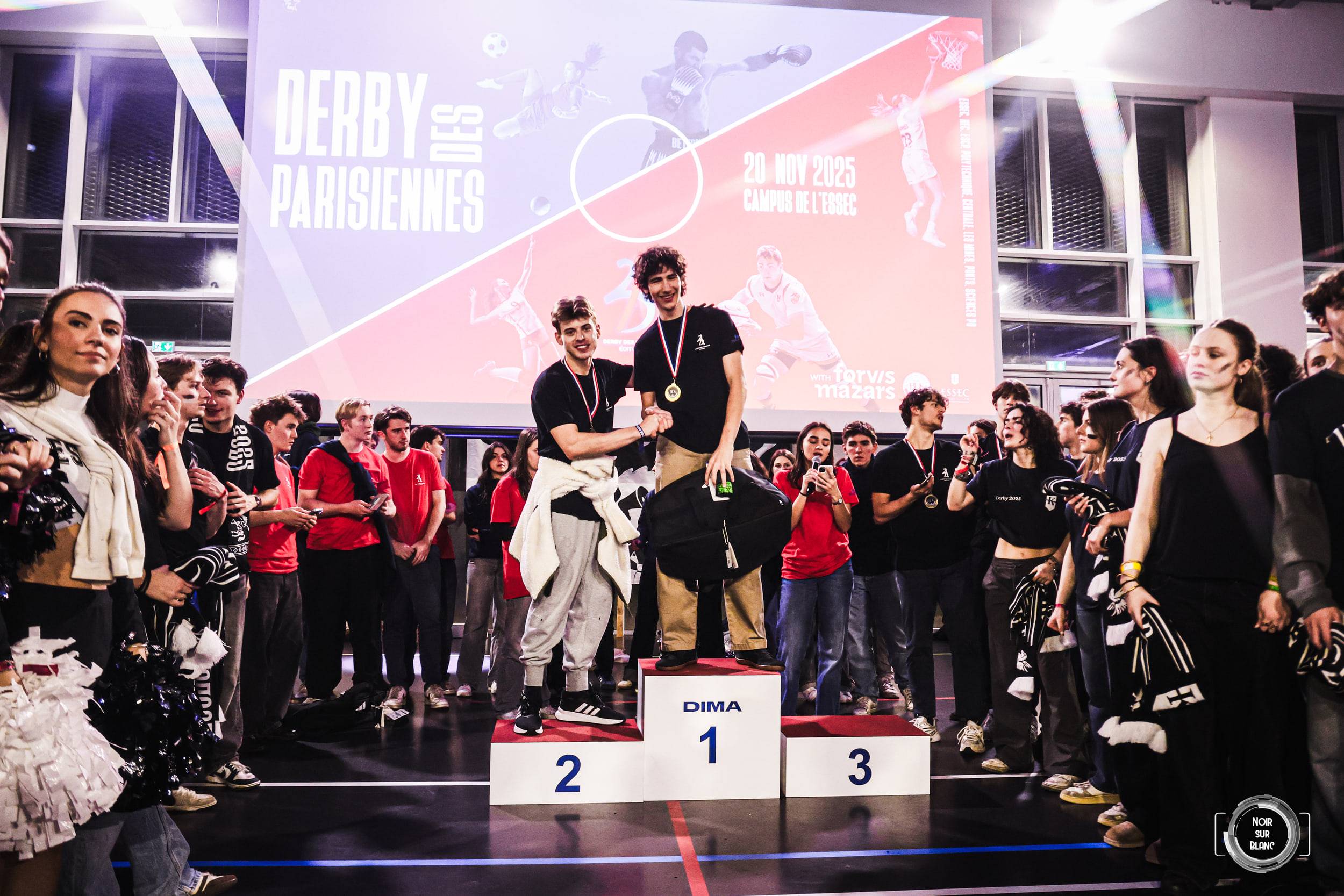 Deux étudiants médaillés lèvent les bras sur la première marche du podium, devant l'écran géant annonçant le Derby des Parisiennes.