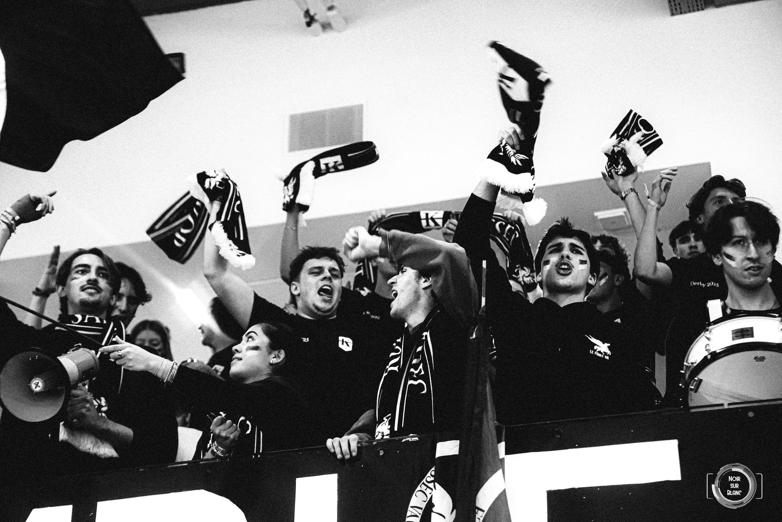 En noir et blanc, les supporters agitent des drapeaux et frappent un tambour au-dessus d'une banderole, mégaphone à la main.