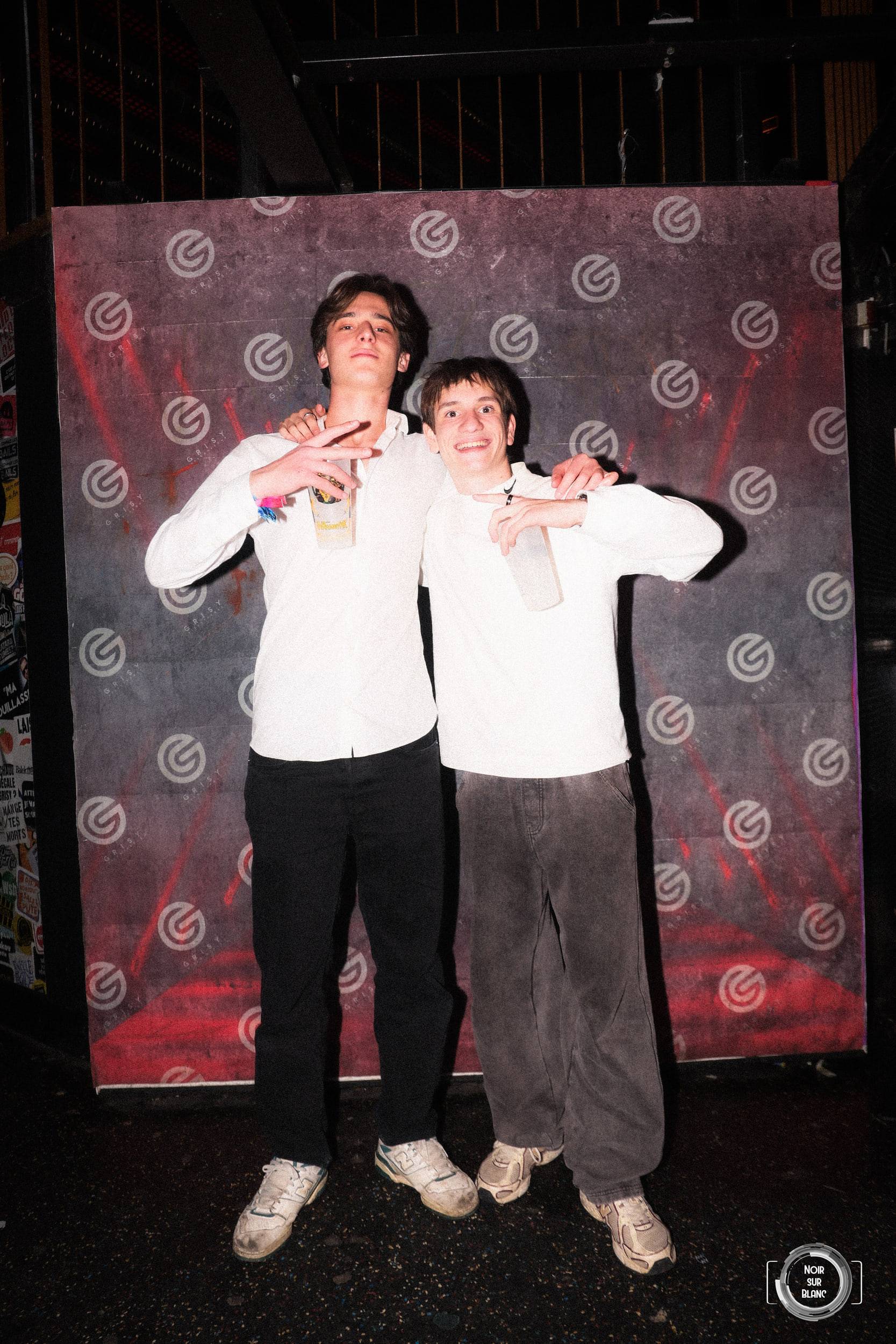 Deux étudiants en chemise blanche posent bras dessus bras dessous devant la toile de fond du photocall, verre à la main.