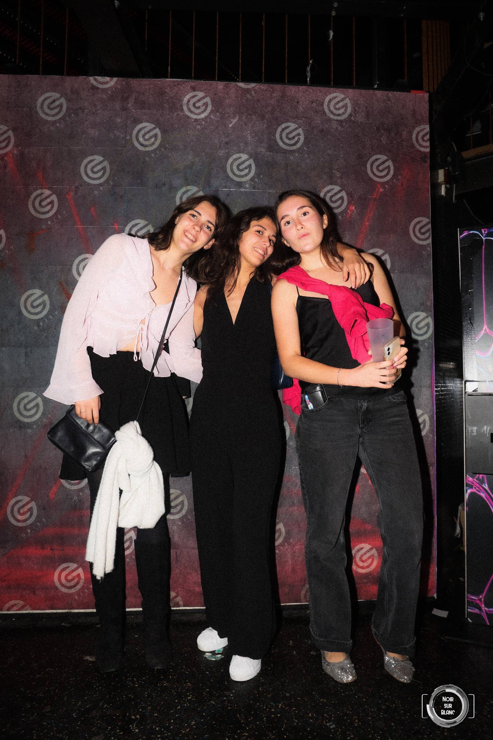 Trois jeunes femmes posent serrées devant la toile de fond du photocall, leur manteau sur le bras.