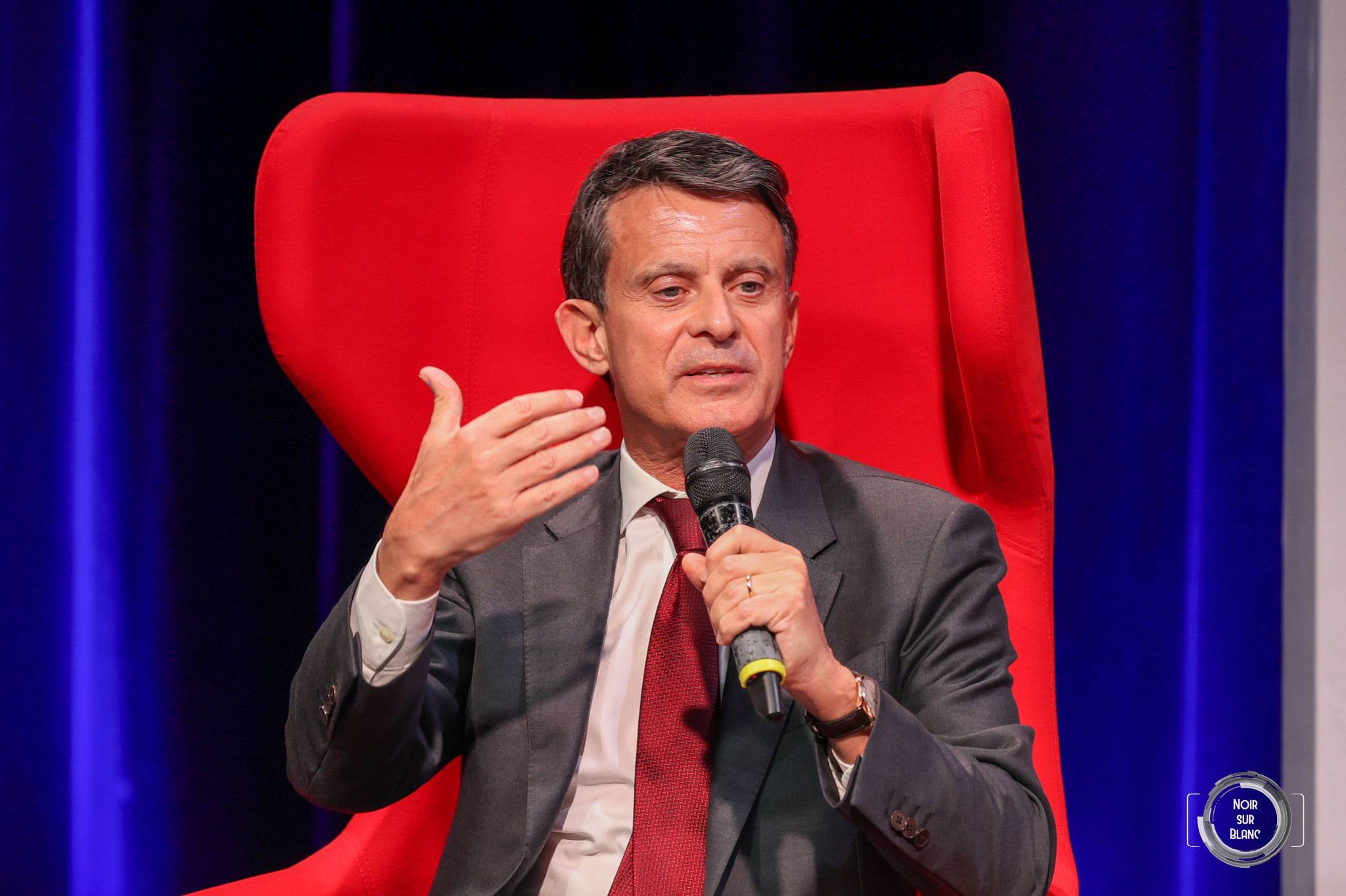 Un intervenant en costume sombre, micro à la main, parle en gesticulant depuis un grand fauteuil rouge.