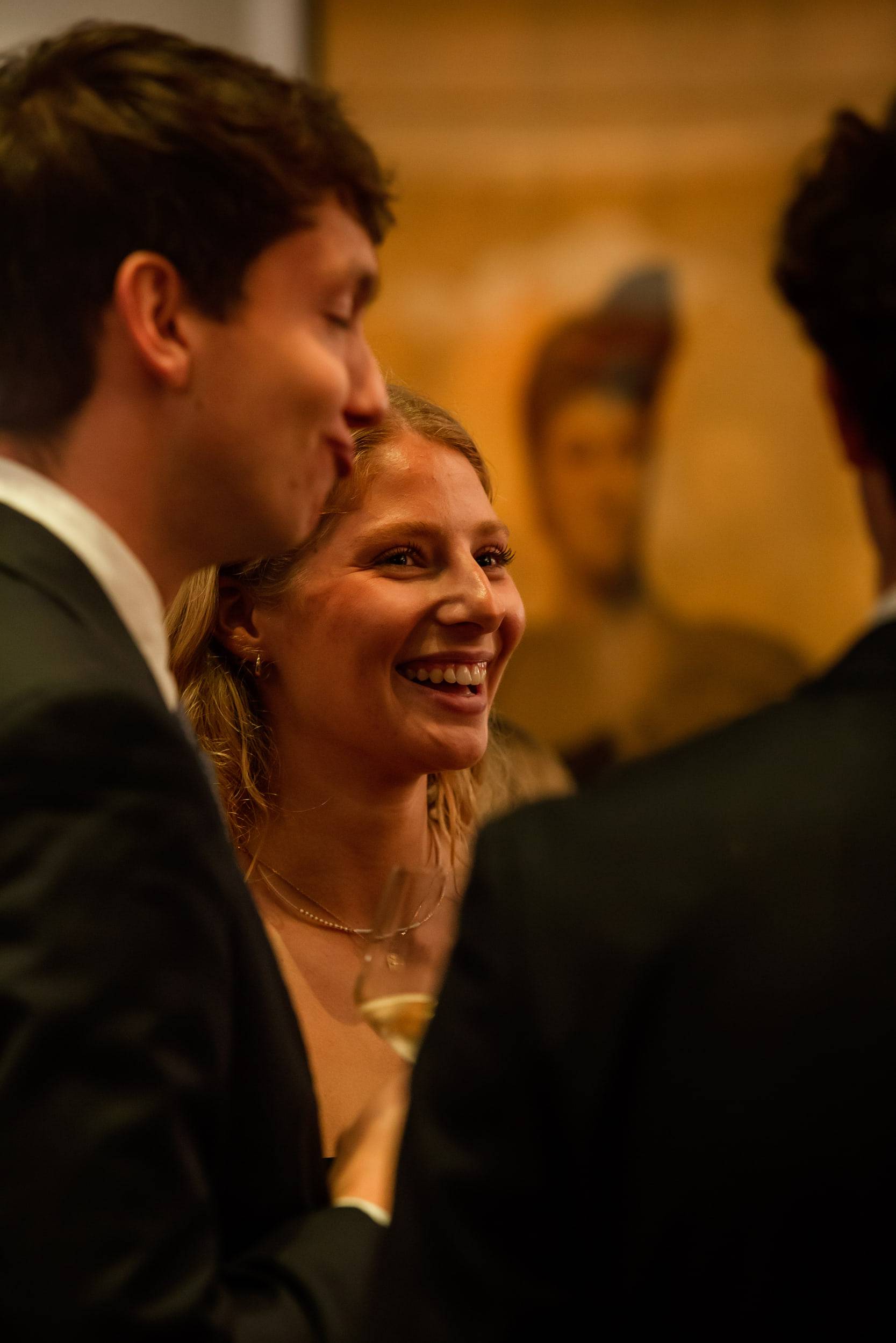 Une étudiante rit aux éclats au milieu d'un groupe en tenue de soirée, devant un tableau ancien.