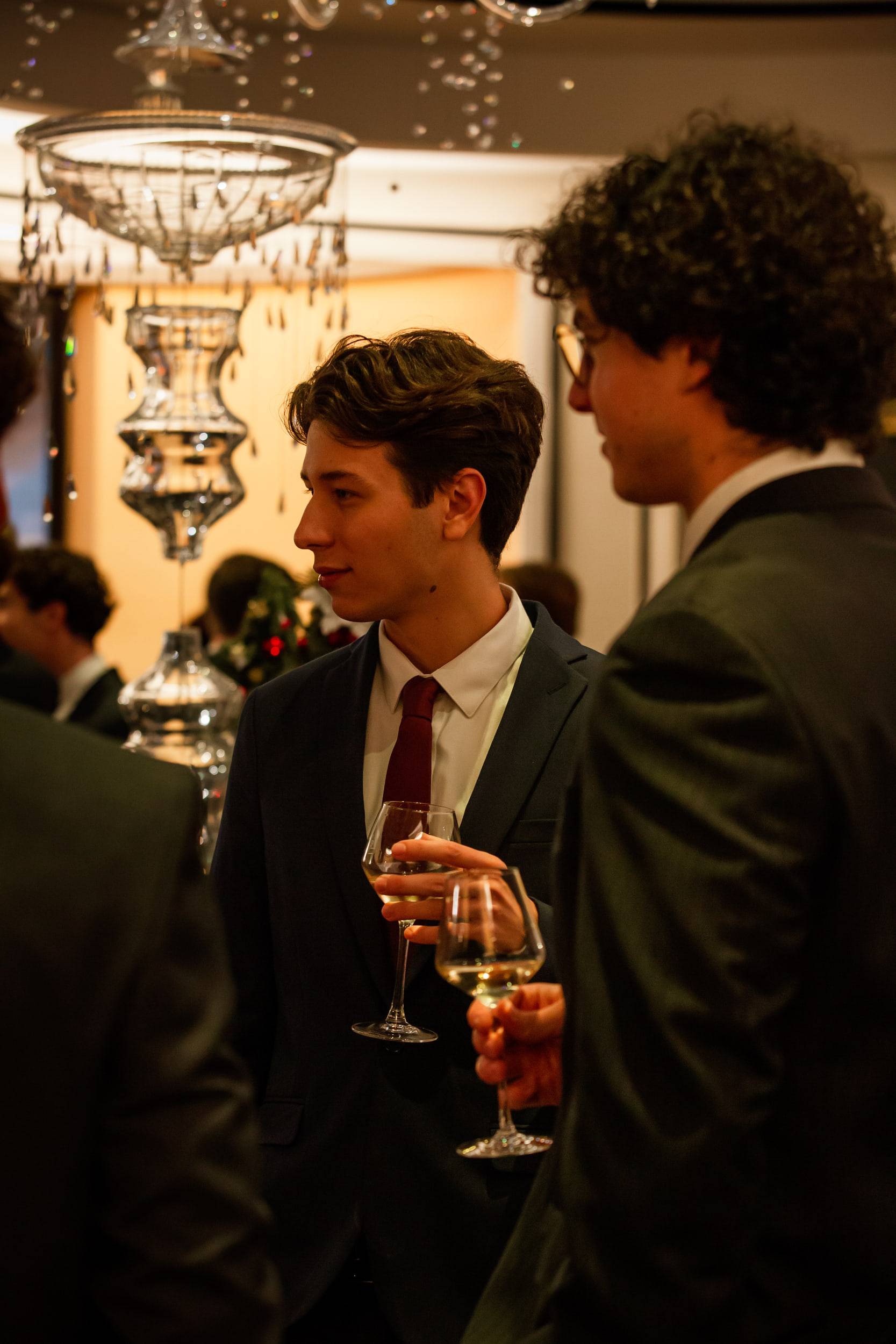 Un étudiant en costume et cravate bordeaux, verre à la main, de profil sous une enfilade de lustres en cristal.