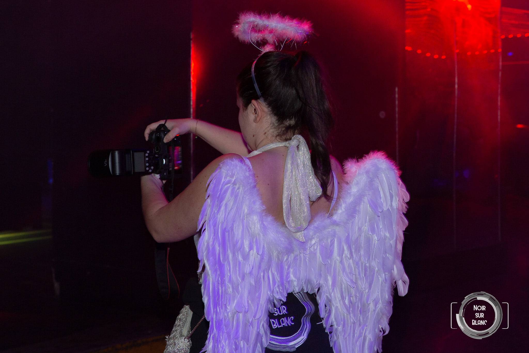 De dos, une fêtarde en grandes ailes d'ange blanches et auréole se fait photographier dans la lumière rouge de la salle.