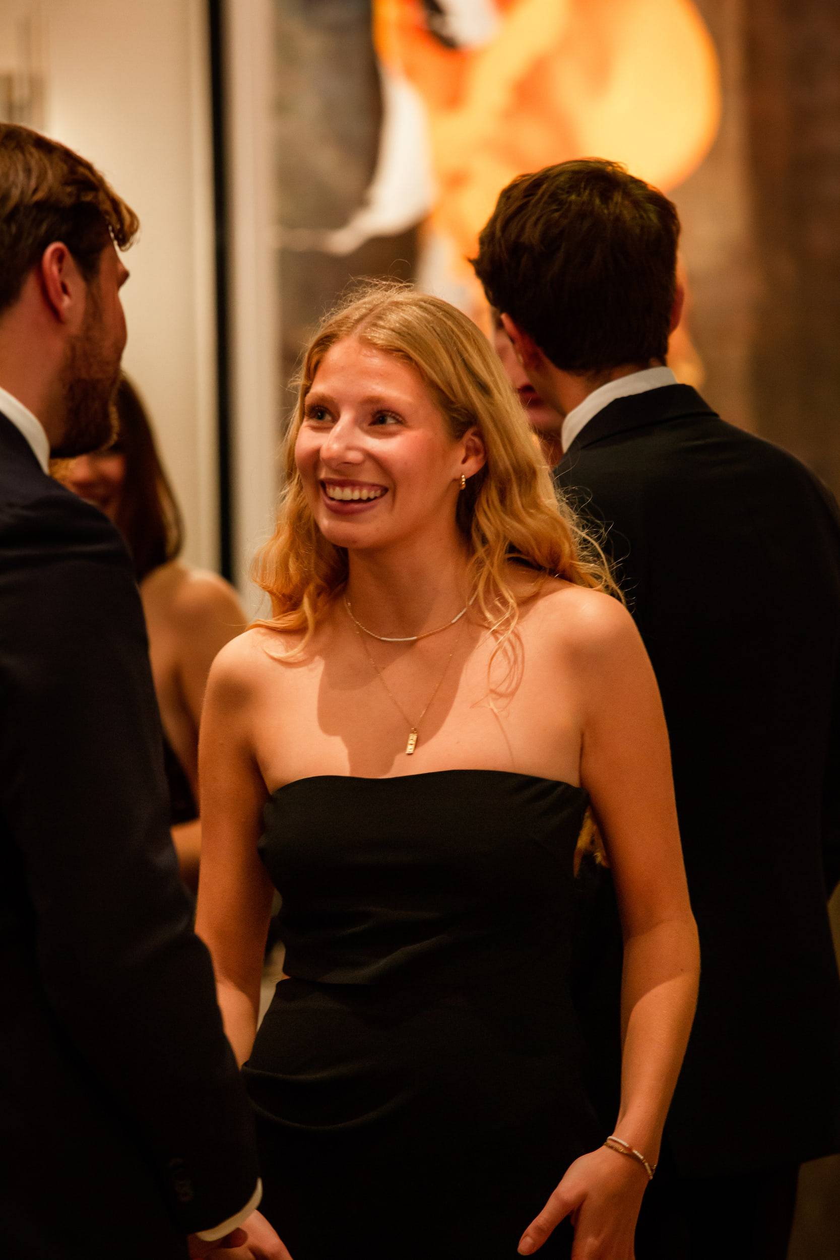 Une étudiante en robe bustier noire sourit, encadrée par deux invités en costume, dans un salon éclairé de bougies.
