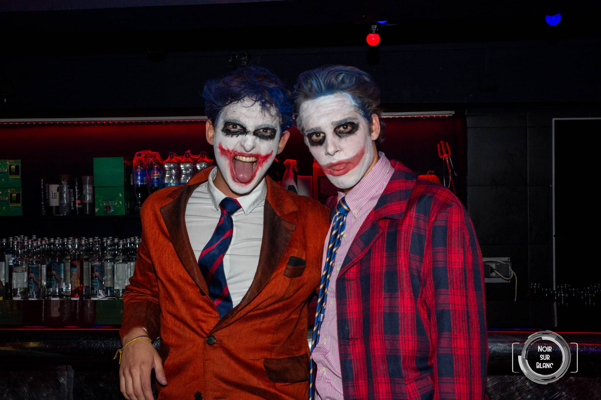 Deux étudiants maquillés en Joker, visage blanc et sourire rouge, posent au bar, l'un en veste orange, l'autre en chemise à carreaux.