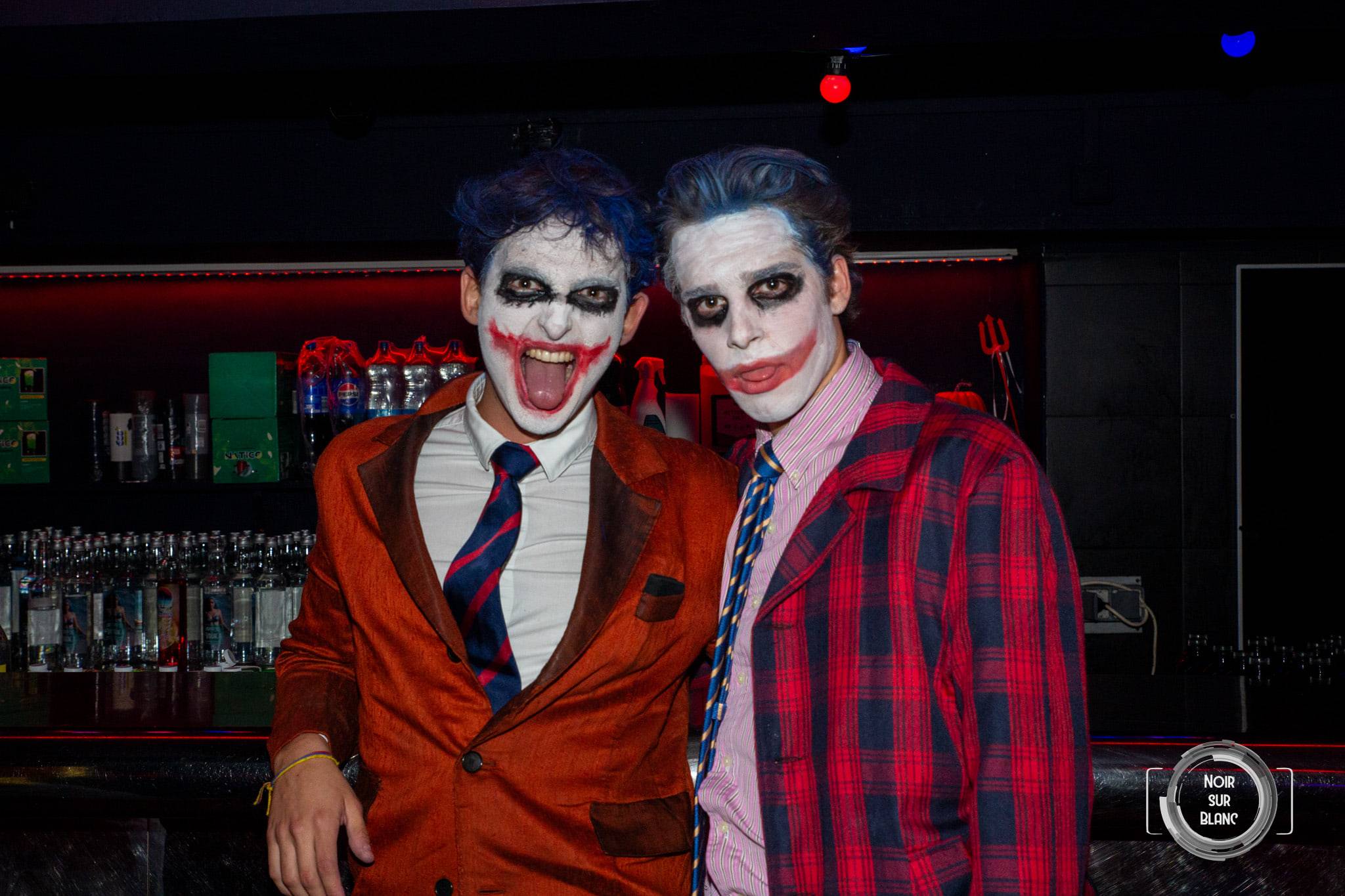 Deux étudiants maquillés en Joker, visage blanc et sourire rouge, posent au bar, l'un en veste orange, l'autre en chemise à carreaux.
