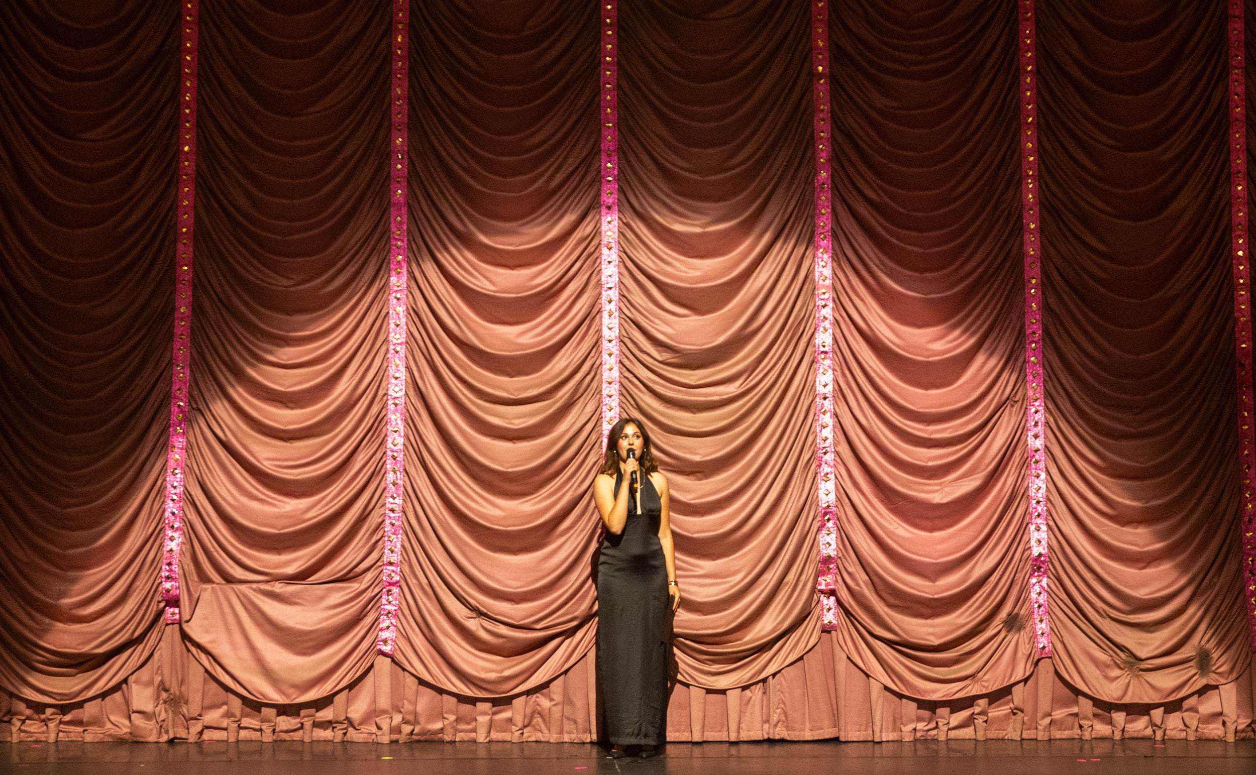 Une chanteuse seule en robe noire, micro à la main, devant l'immense rideau drapé rose du théâtre.