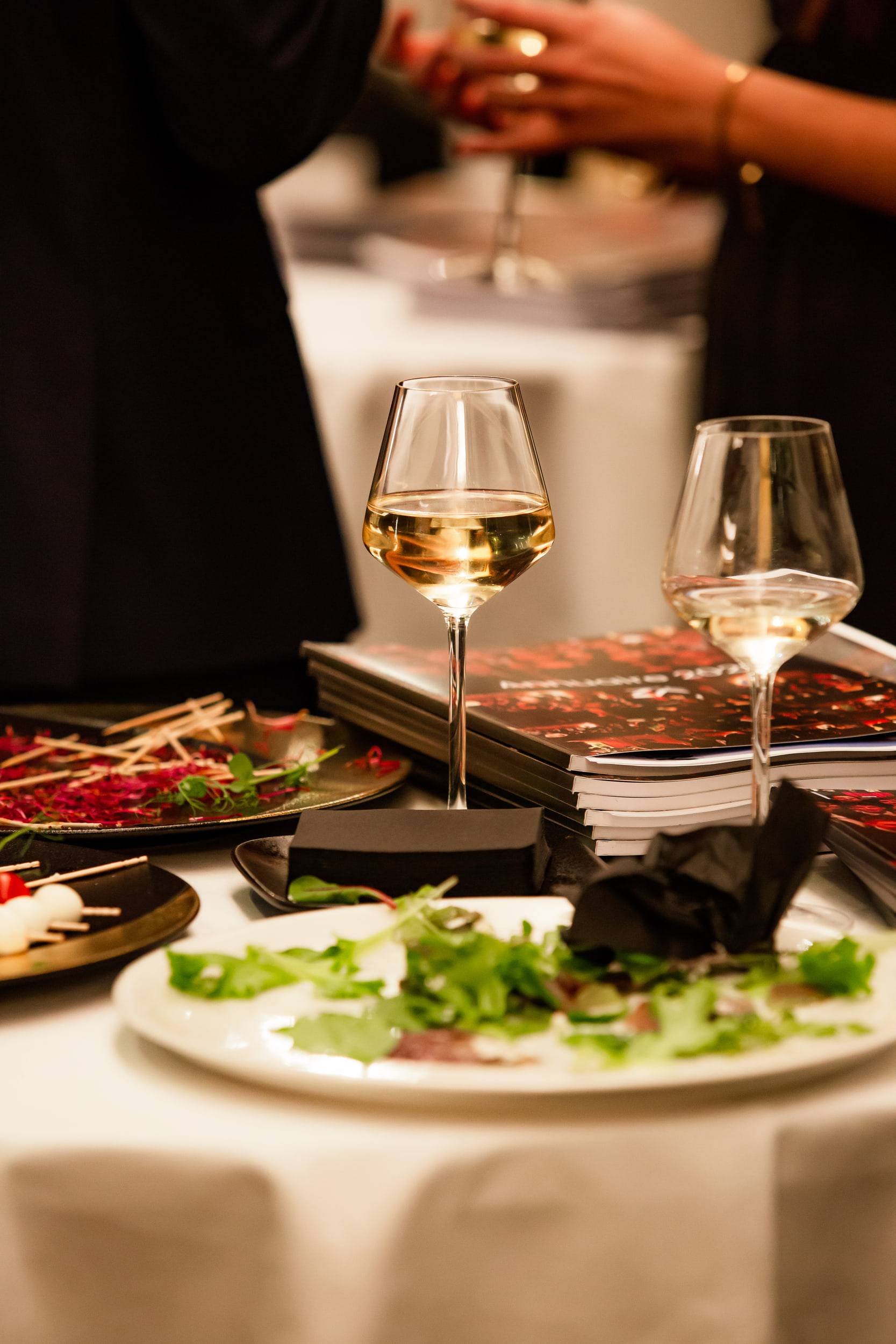 Sur une nappe blanche, deux verres de vin et une assiette de charcuterie et de salade, dans la lumière chaude du salon.