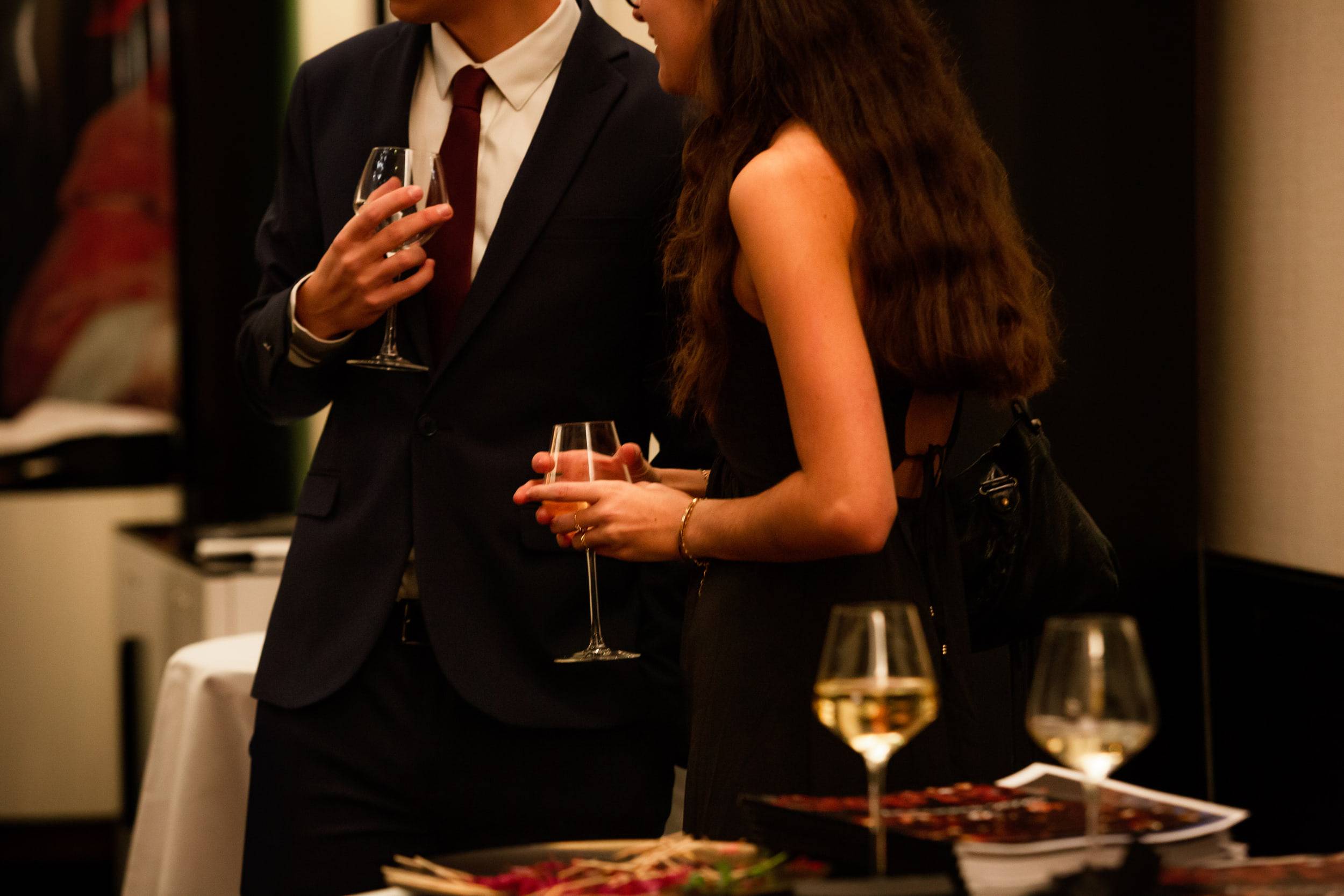 Un étudiant en costume et une étudiante en robe noire discutent, verres à la main, près d'un buffet.
