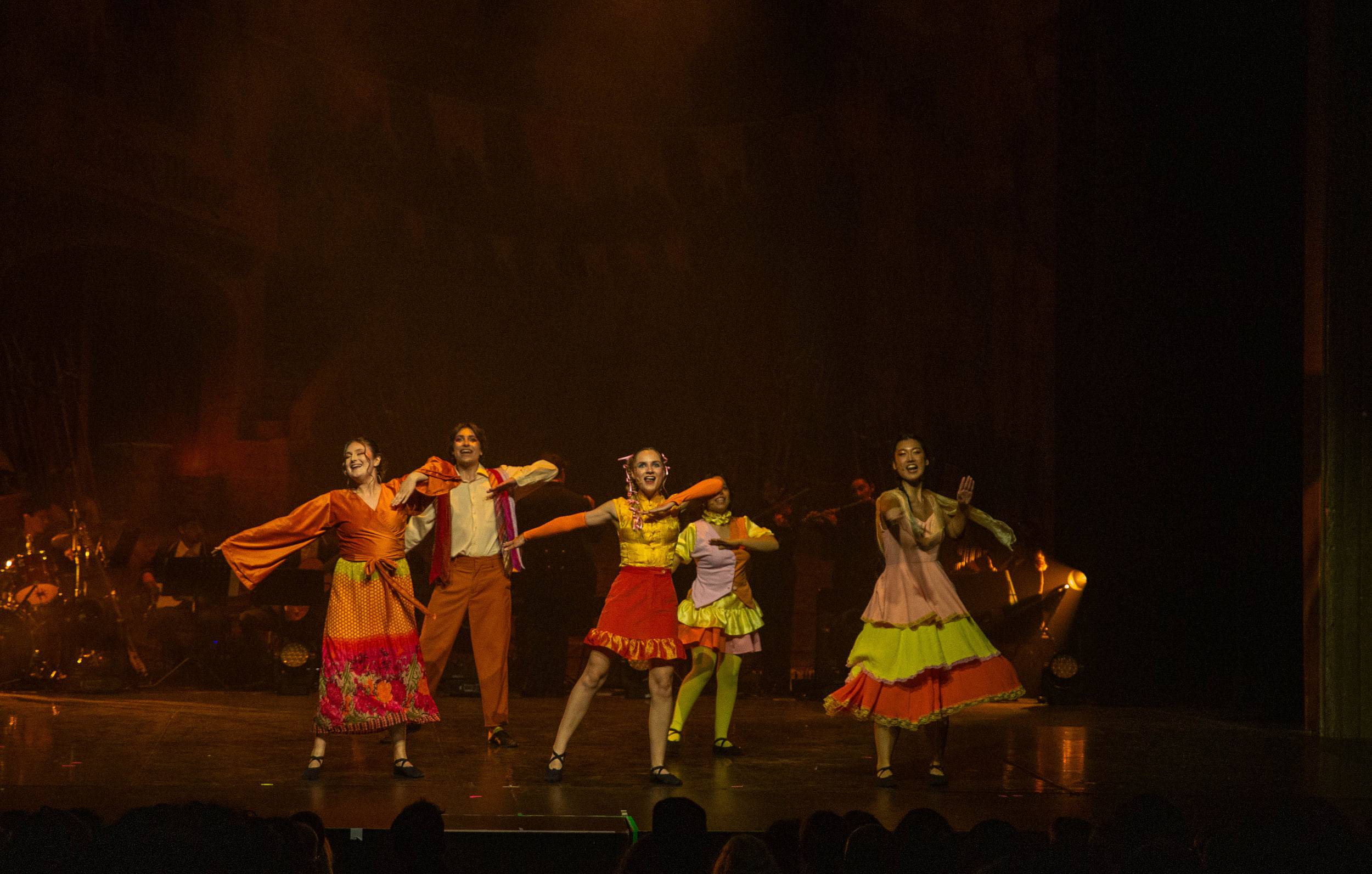 Cinq danseurs en costumes orange et jaunes, bras écartés, sur un plateau éclairé de lumière chaude.