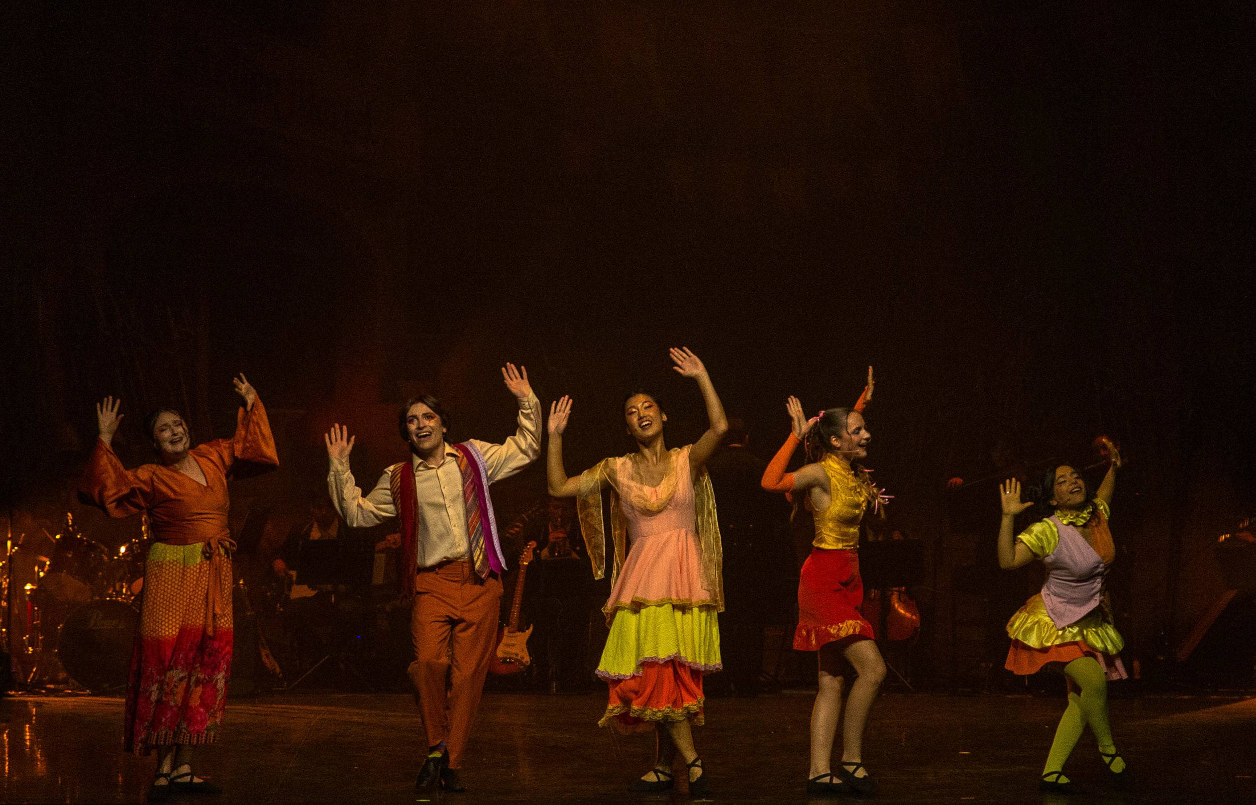 Six comédiens en costumes colorés lèvent les bras dans un même geste, les musiciens en fond de scène.