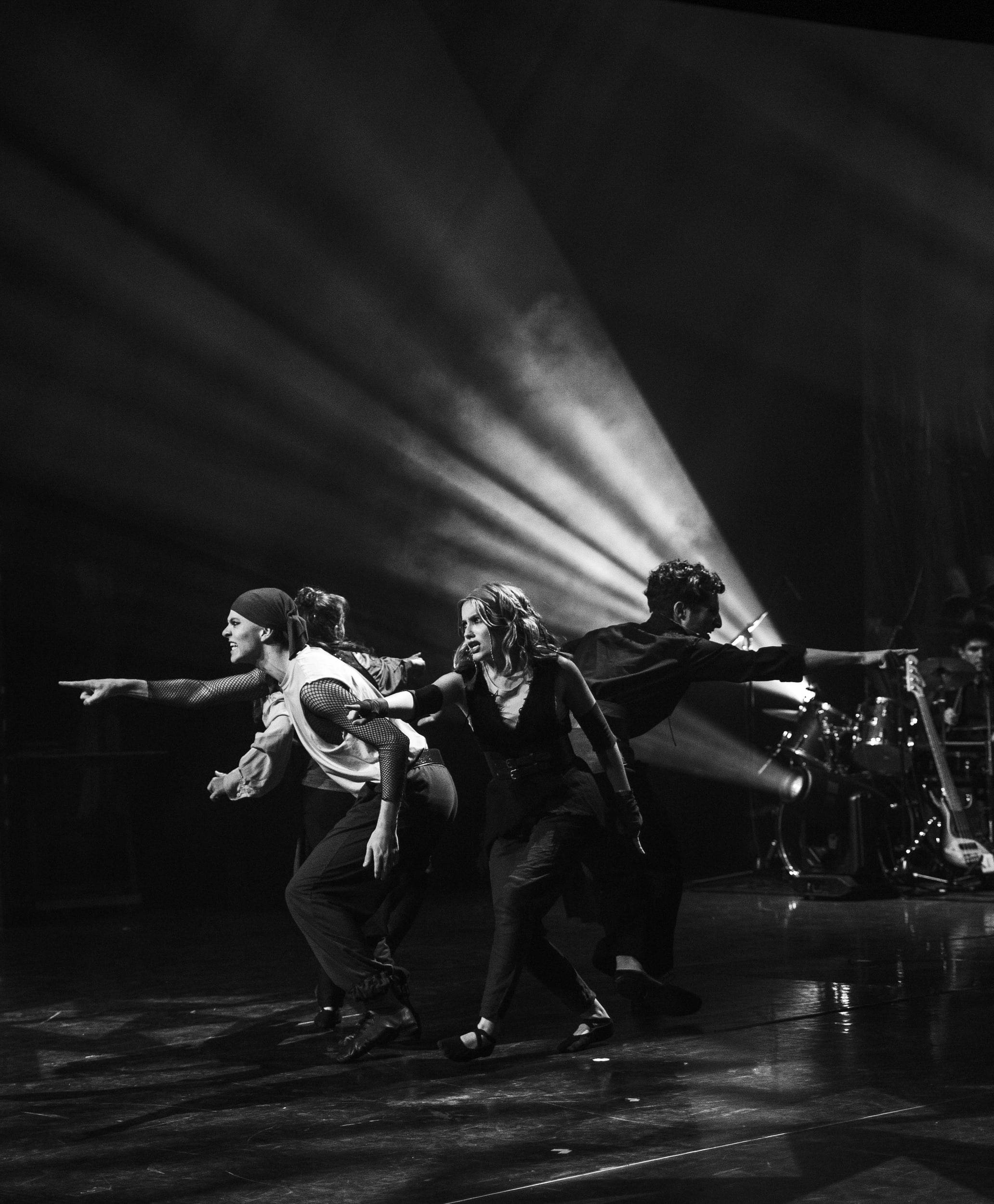 Photographie en noir et blanc : quatre danseurs élancés, bras tendus, traversés par les faisceaux d'un projecteur.