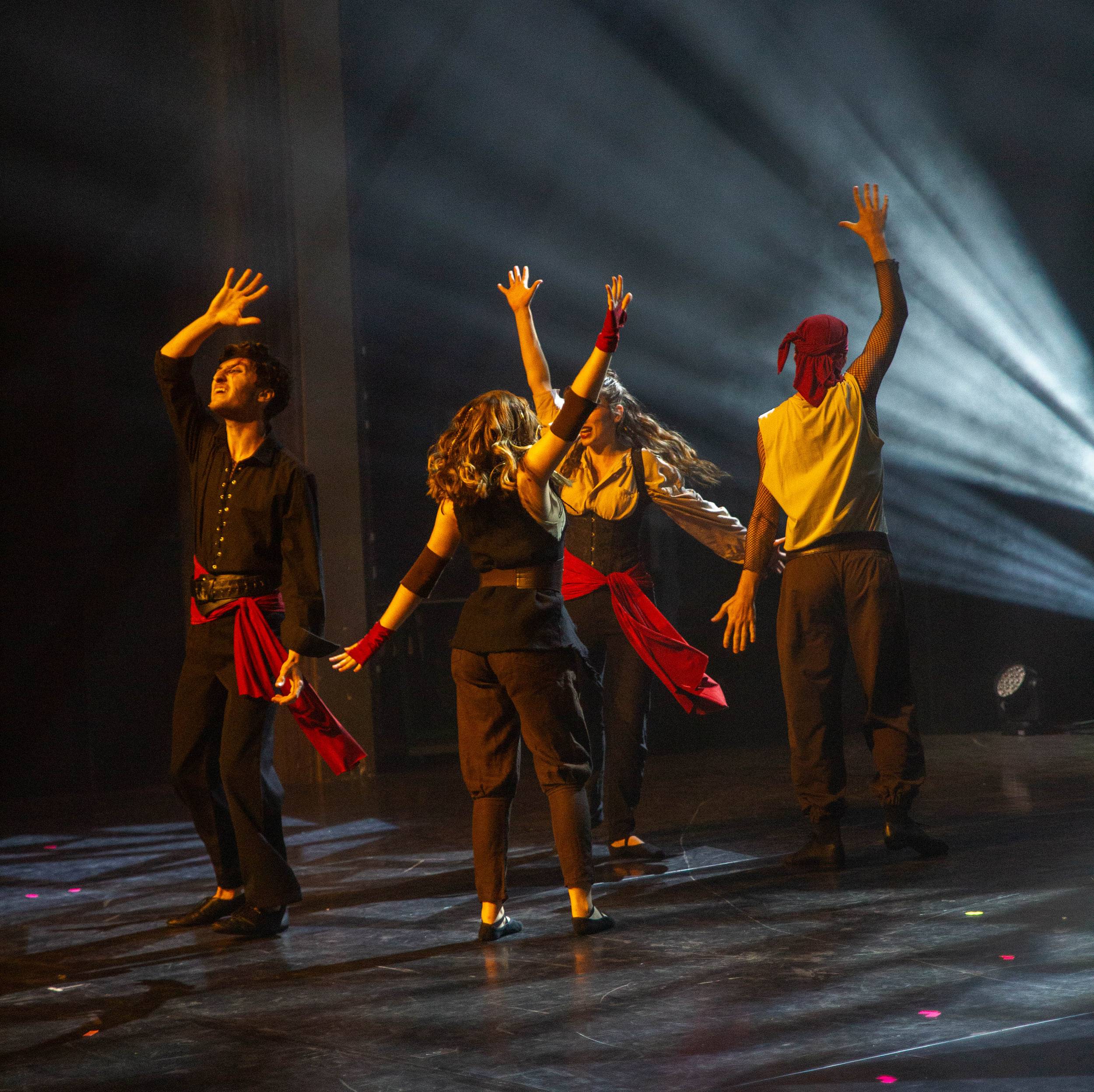 Quatre danseurs en chemises sombres et ceintures rouges, bras levés, dans un rai de lumière bleue.