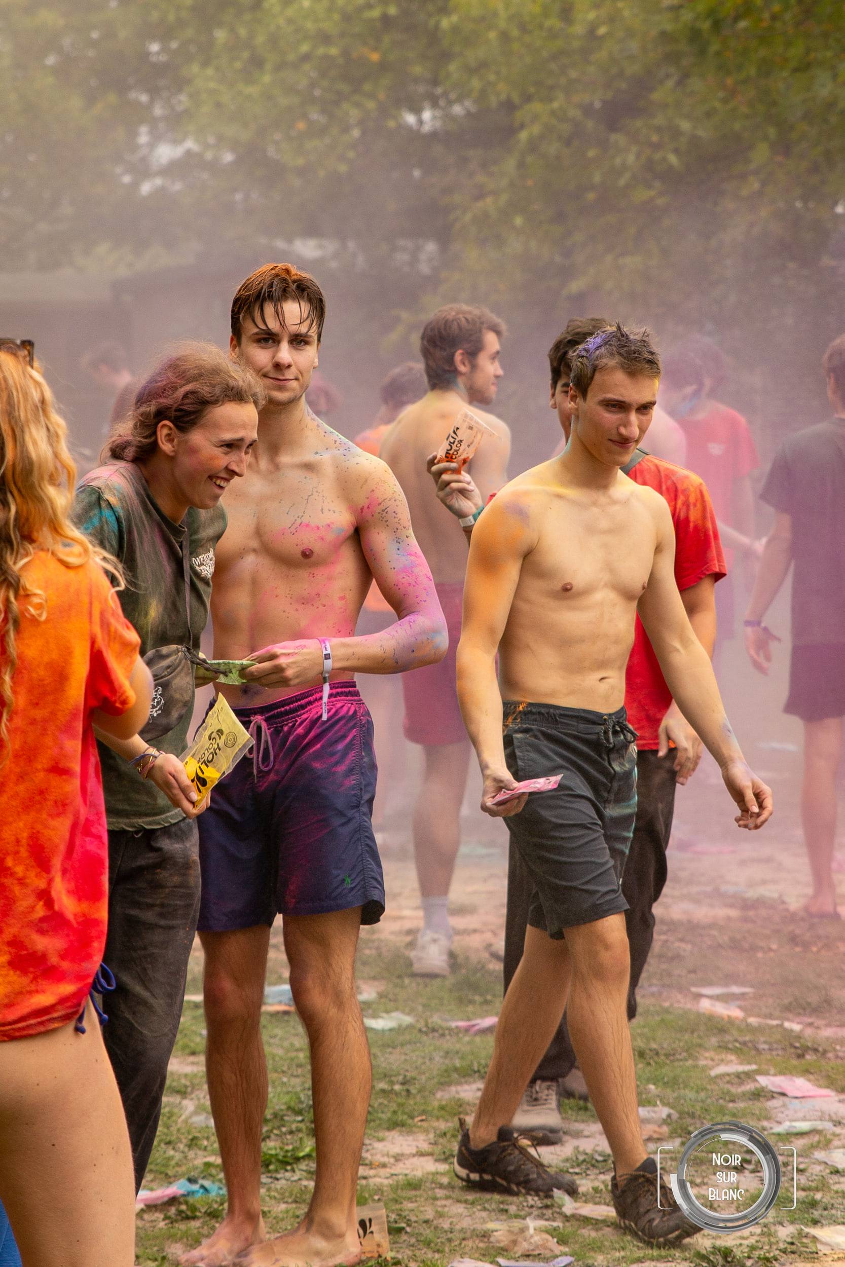 Dans un nuage de pigment en suspension, des étudiants torse nu et maculés de rose et de violet tiennent des sachets de poudre, à la lisière d'un bois.