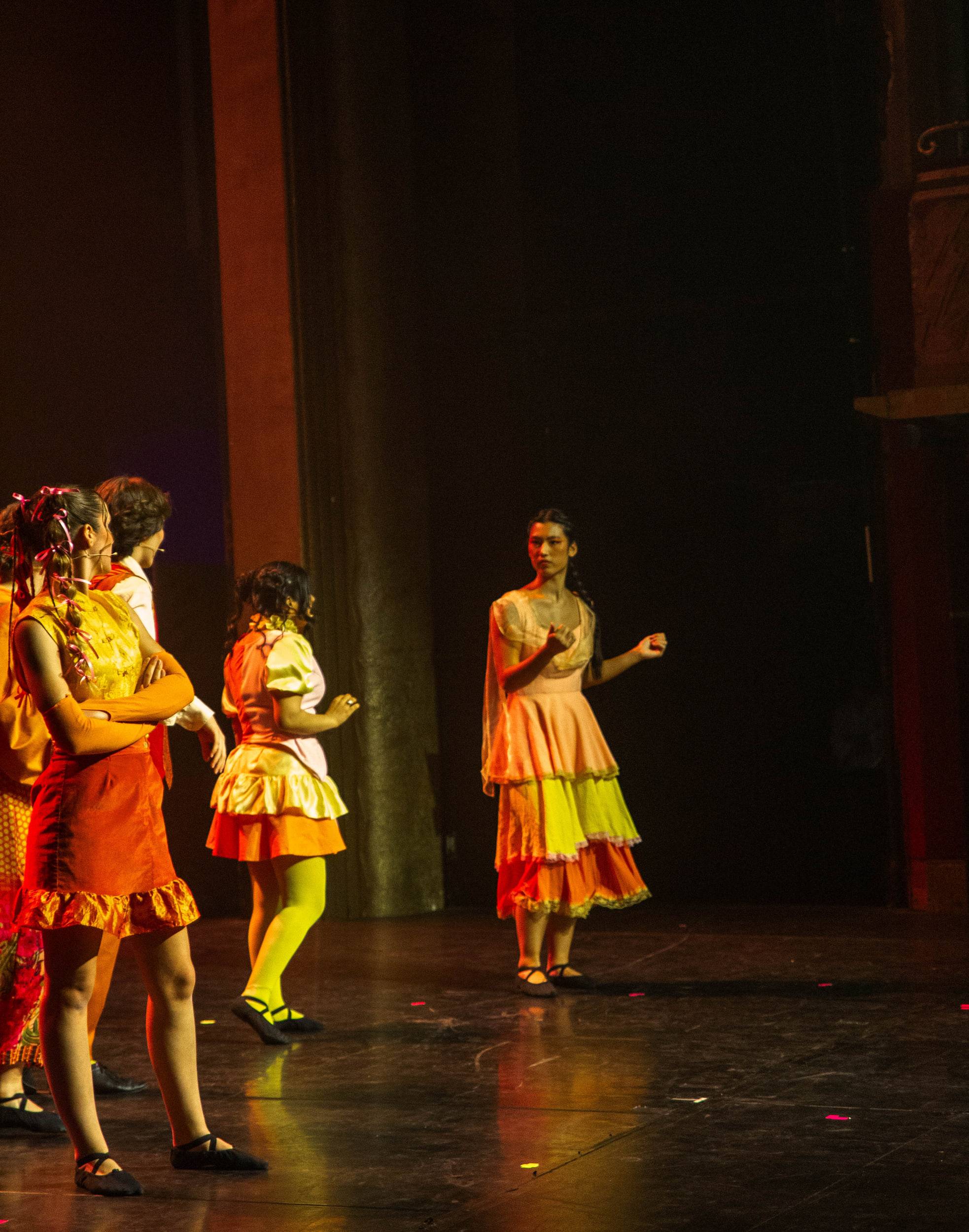 Un groupe de comédiennes en robes orange et jaune à volants se tourne vers l'une d'elles, sur un plateau doré.