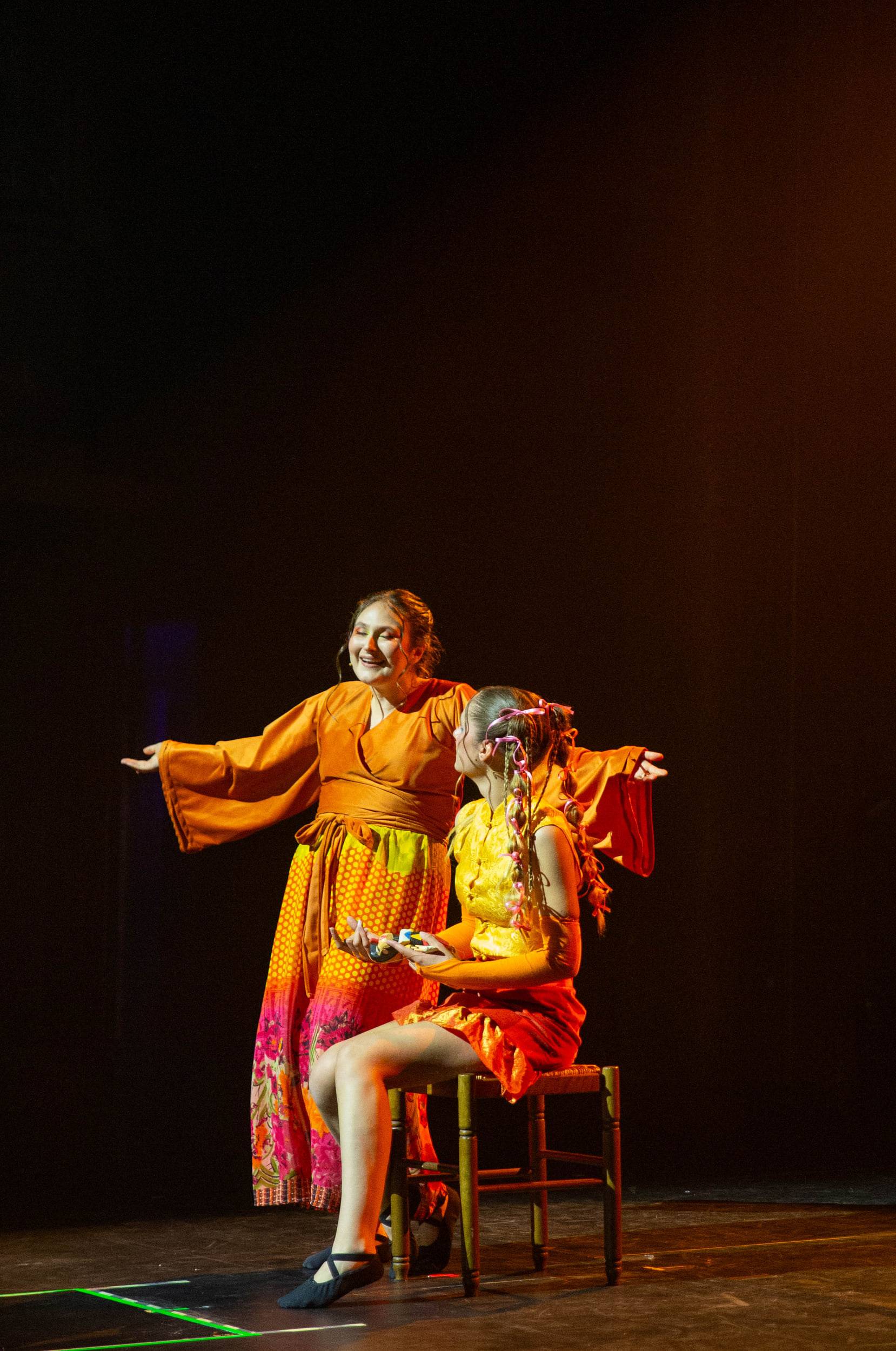 Deux comédiennes en orange, l'une assise sur une chaise, l'autre debout derrière elle, bras ouverts.