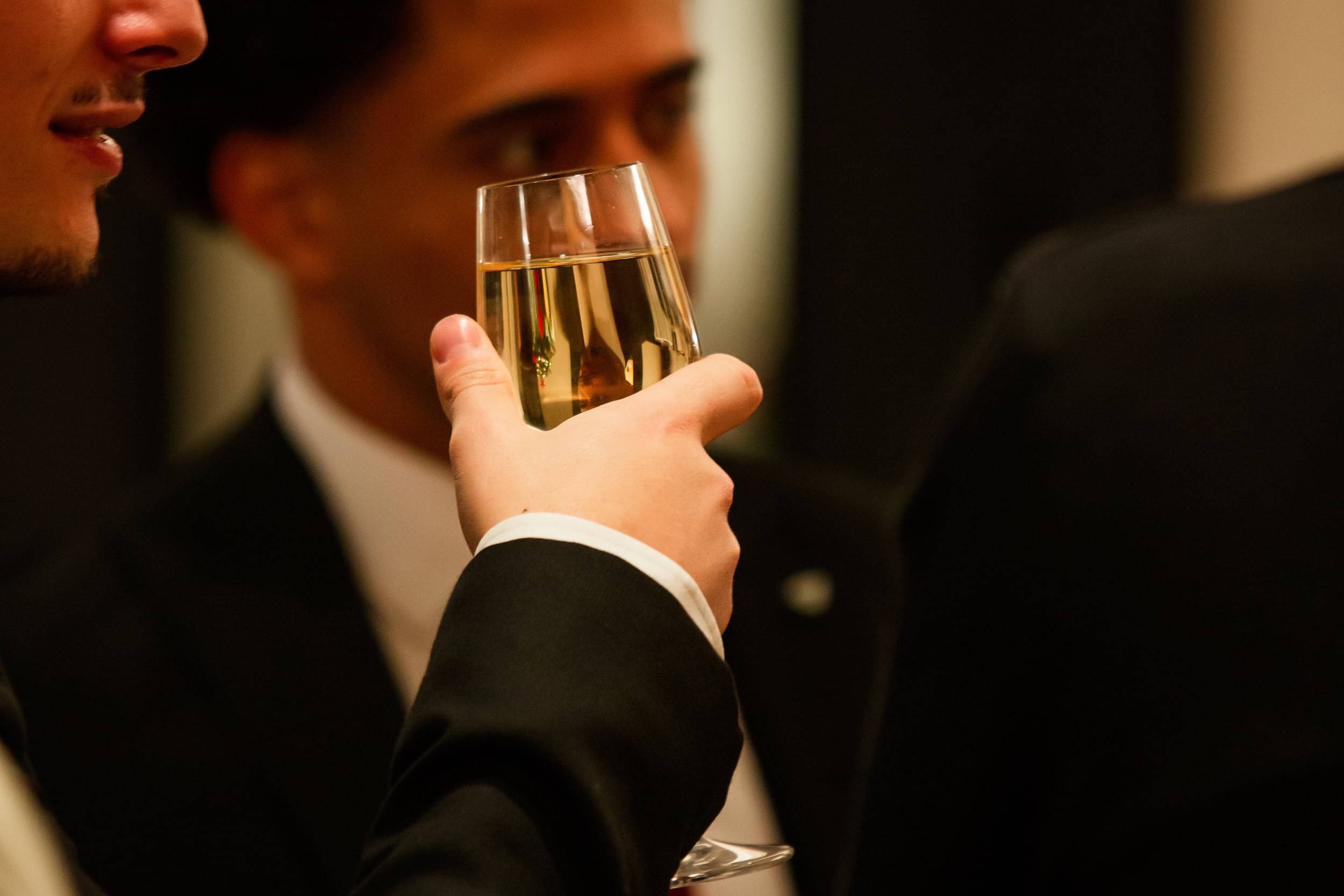 Une main lève une coupe de champagne au premier plan, tandis que deux invités en costume trinquent dans le flou derrière.