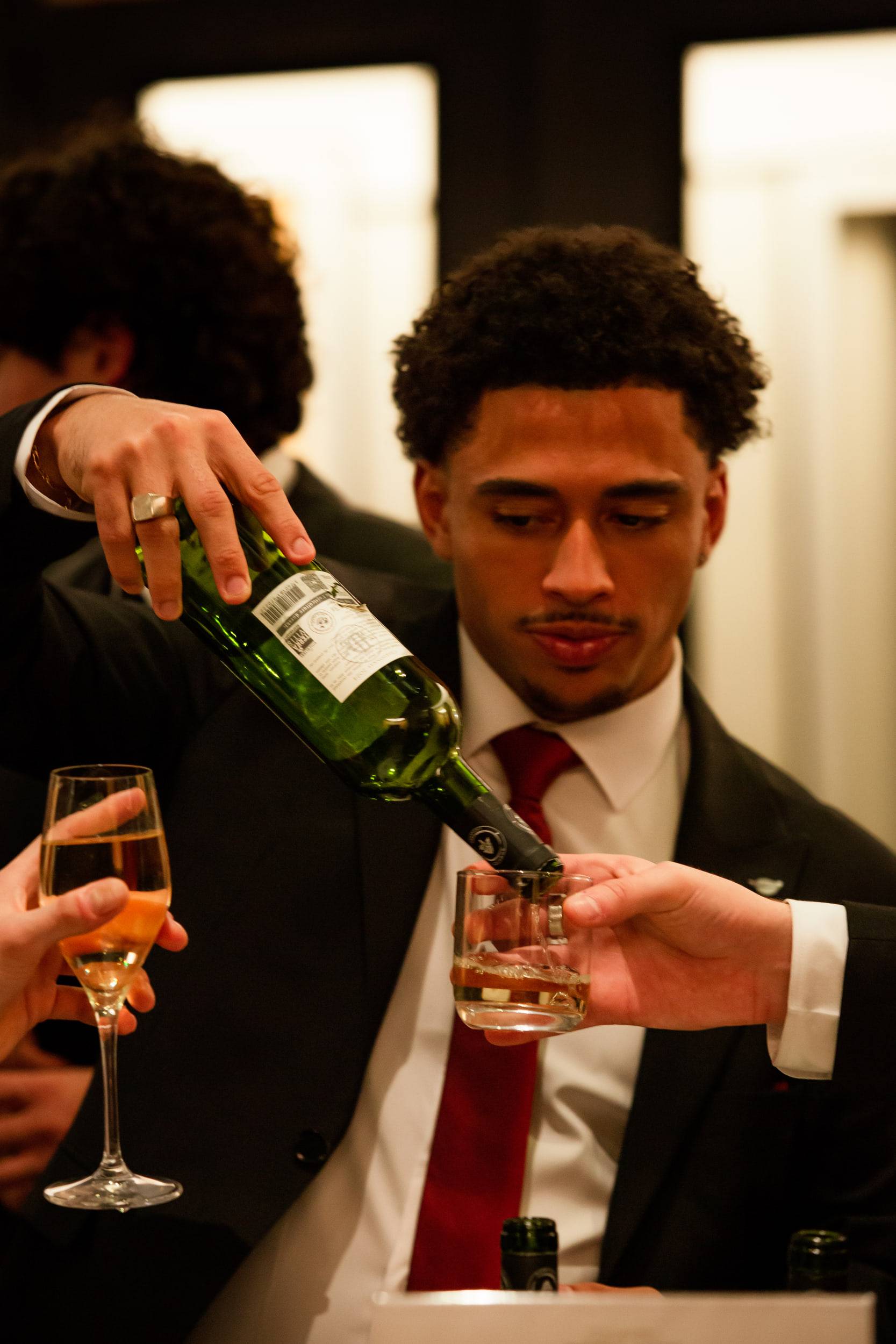 Un étudiant en costume et cravate rouge verse du vin blanc dans le verre tendu d'un invité.