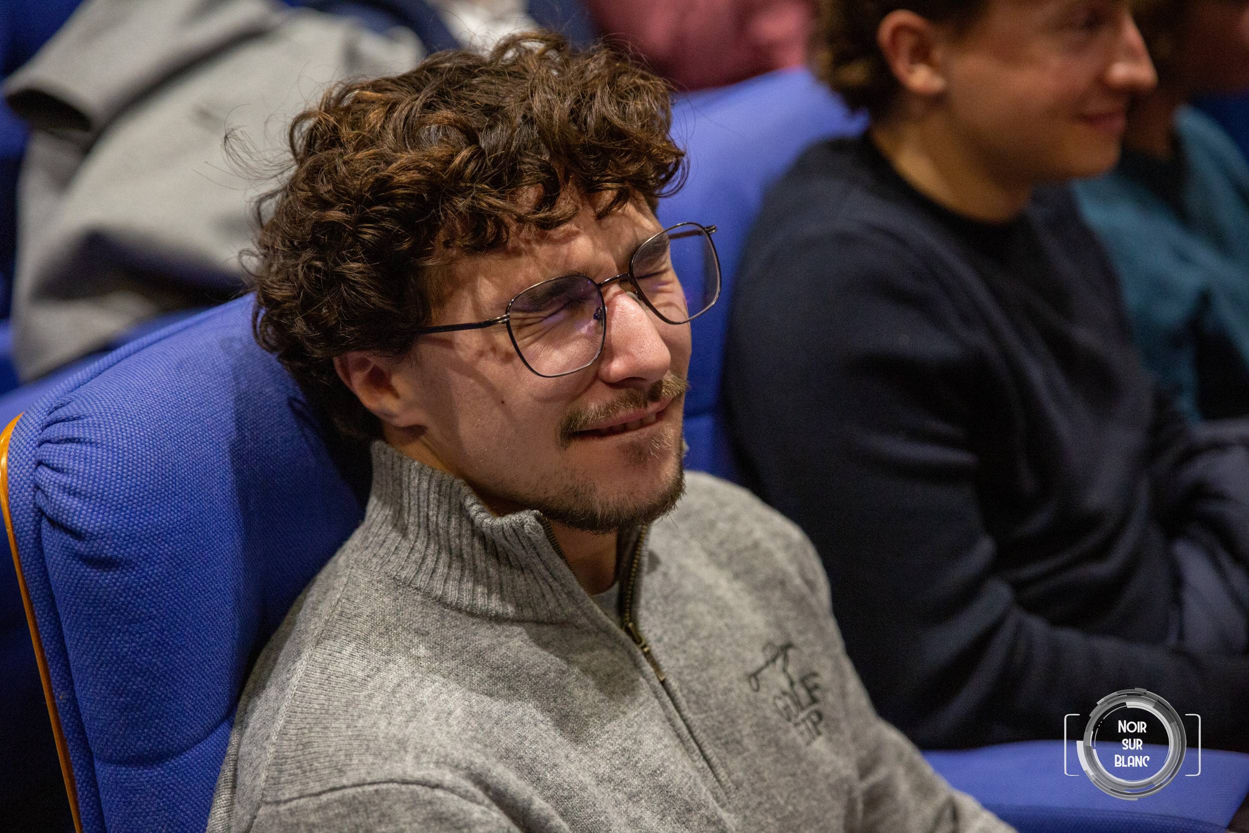 Portrait d'un spectateur à lunettes, en pull gris, souriant dans un fauteuil bleu au milieu de l'assistance.