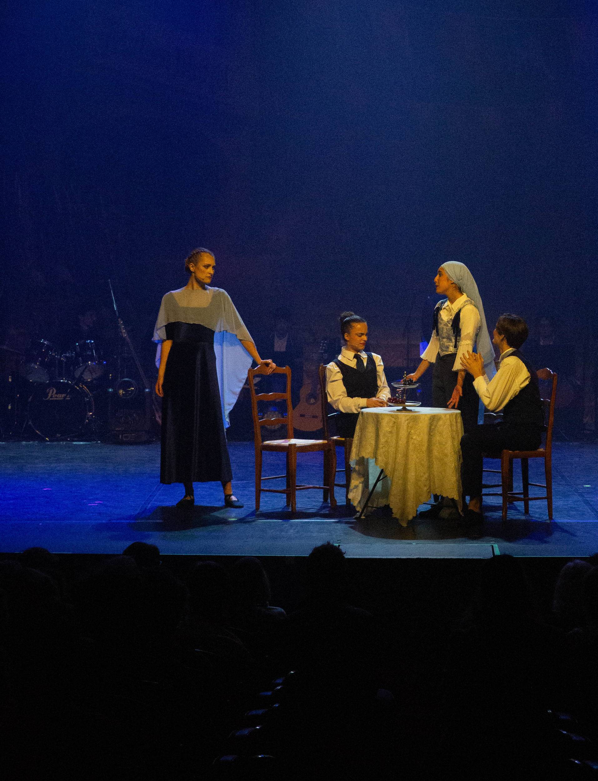 Quatre comédiens autour d'une table dressée, une femme debout à gauche, dans une lumière bleue.