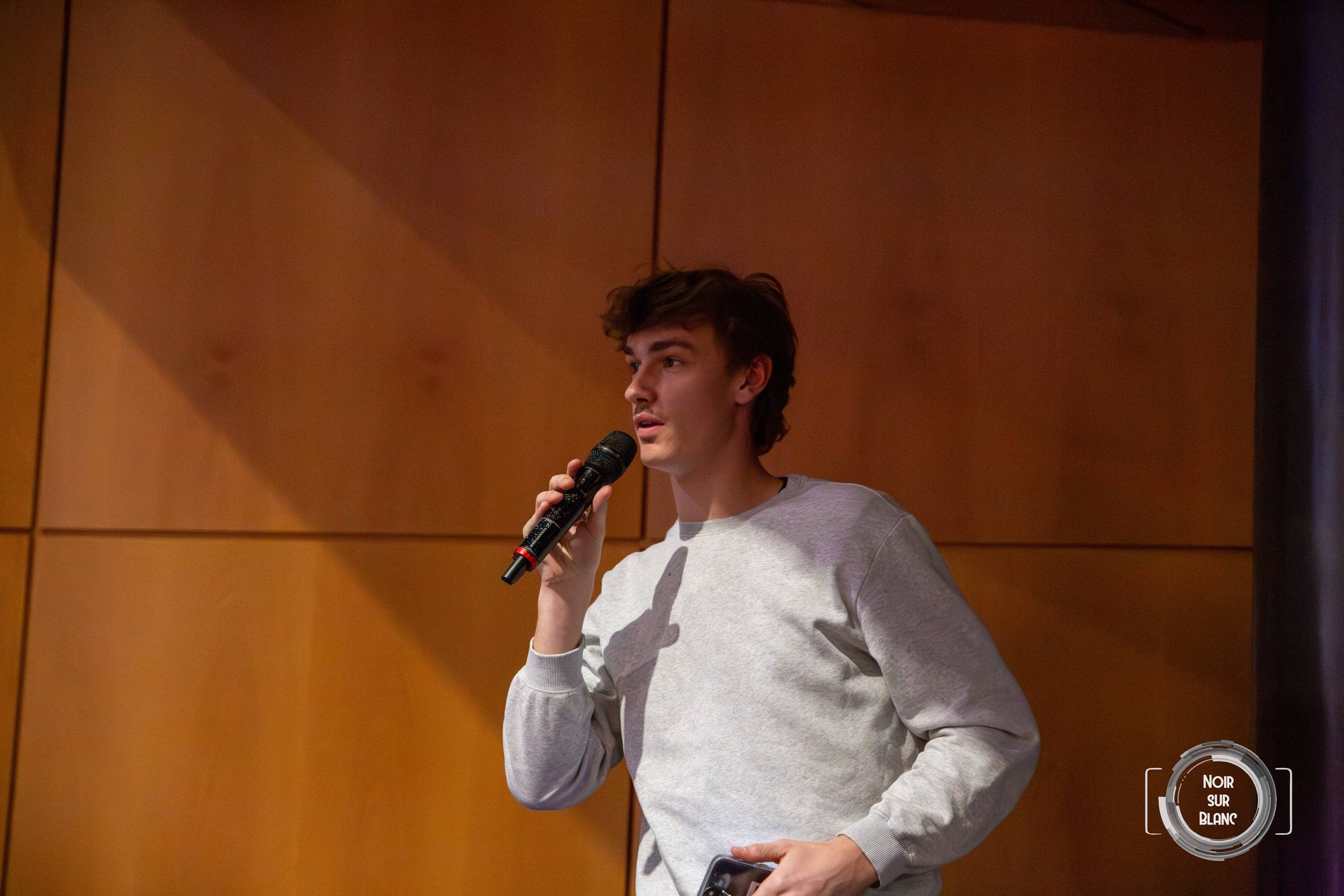 Un étudiant debout, micro à la main, pose sa question devant un mur de bois clair.