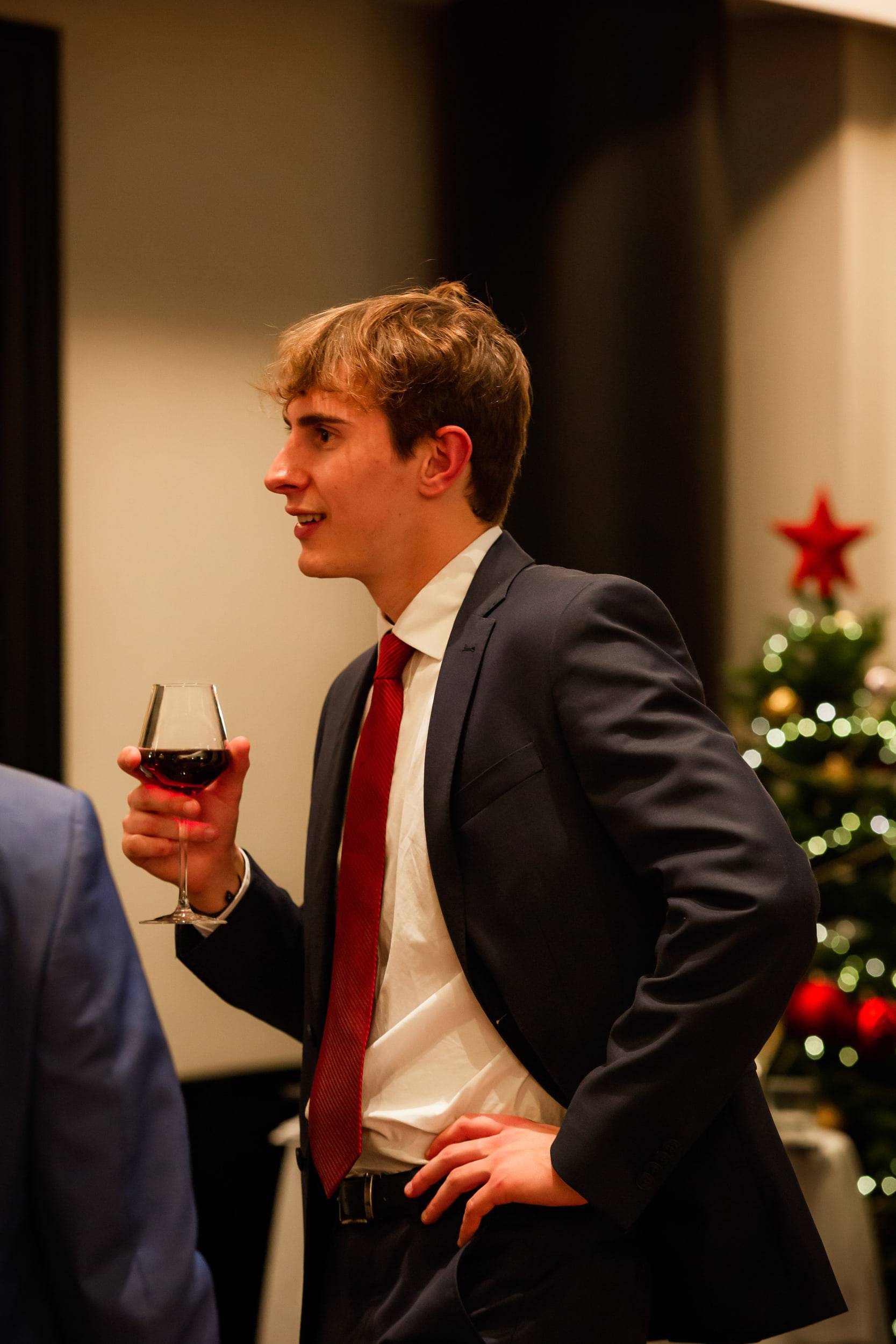 Un étudiant en costume sombre et cravate rouge, verre de vin à la main, devant un sapin de Noël.